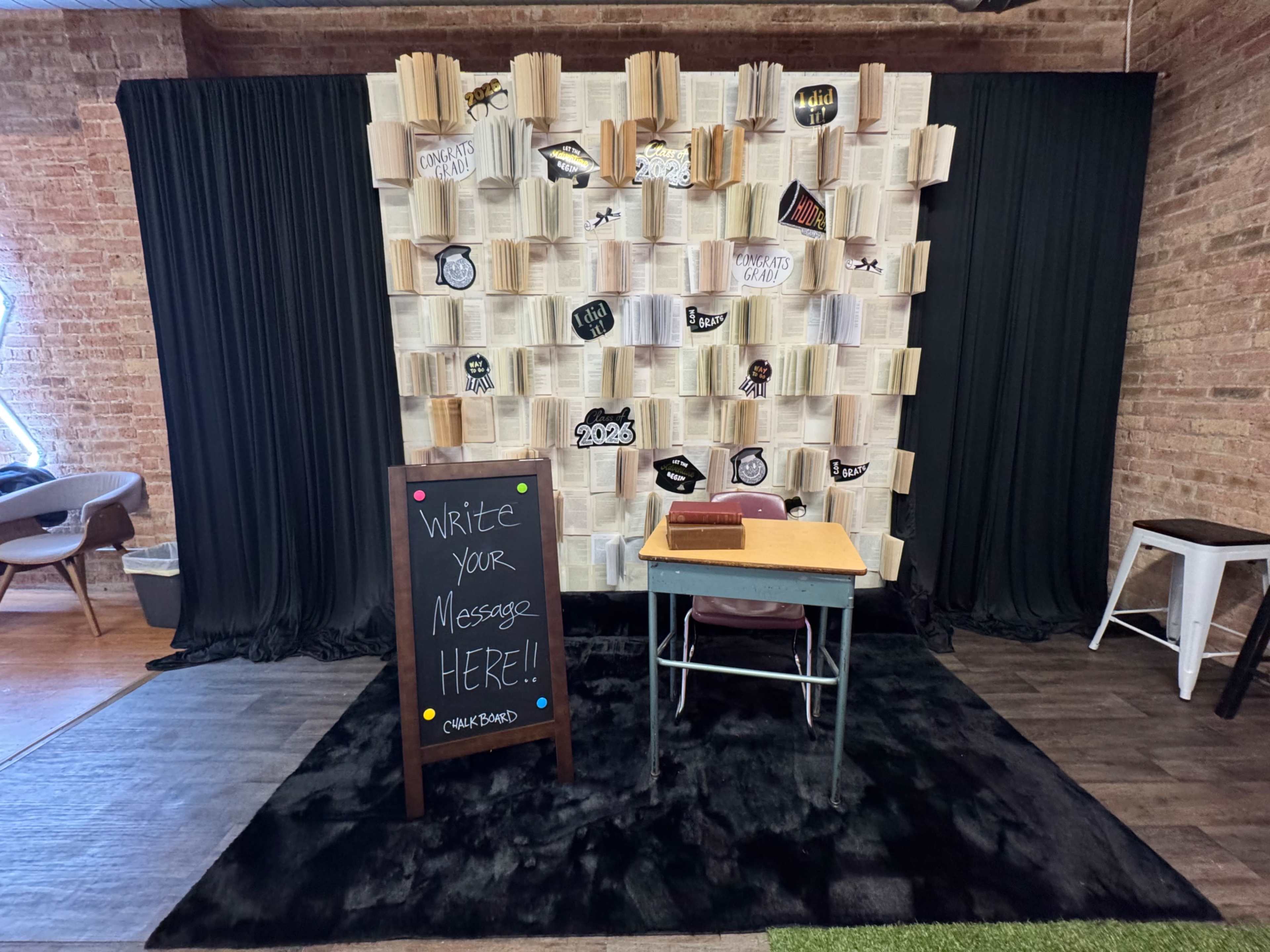 The image shows a decorated wall made of book pages, with a small desk and a chalkboard in front inviting people to write messages.