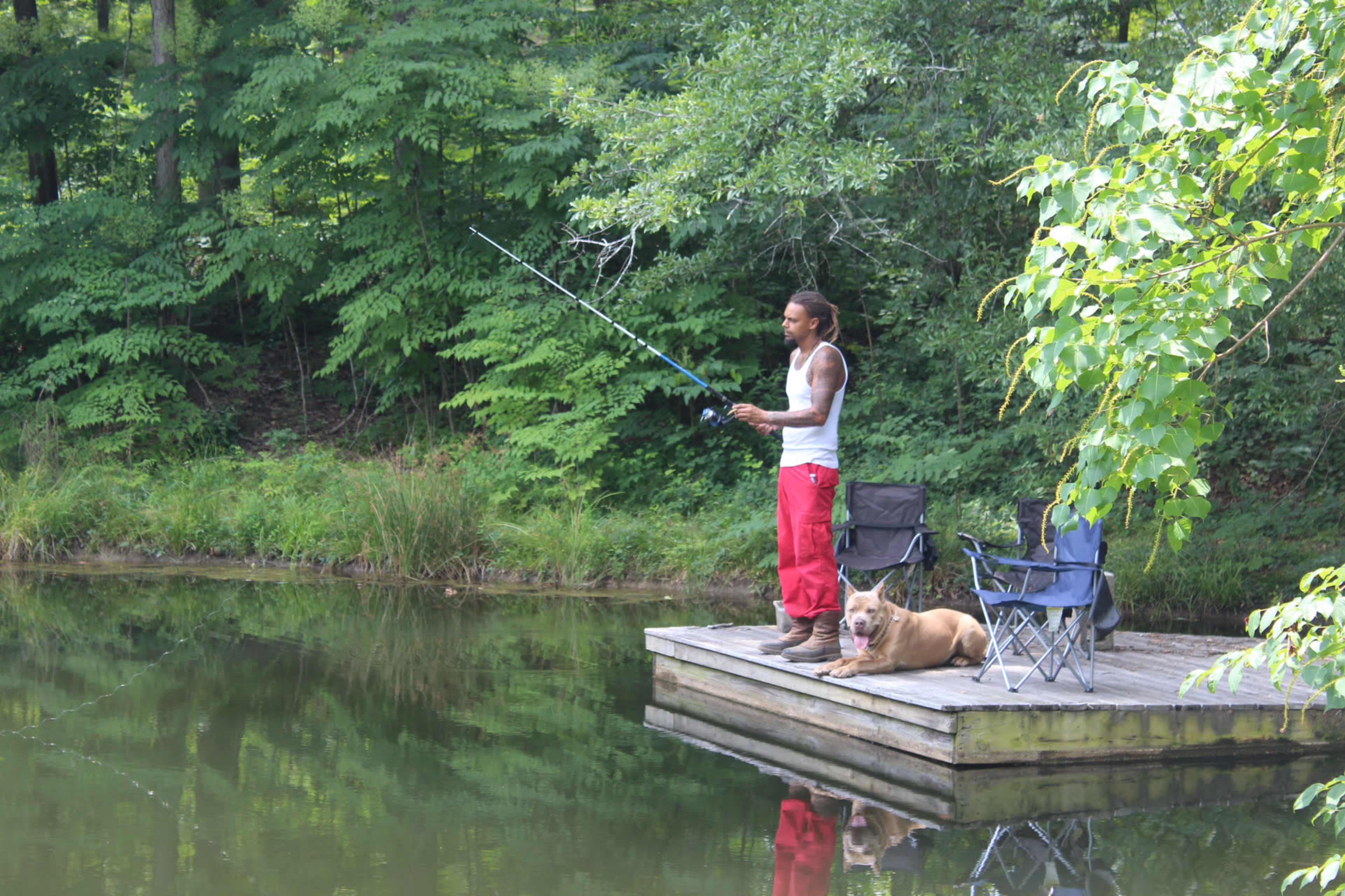 A person stands on a wooden dock fishing beside a dog lying on the edge of a pond, surrounded by dense greenery.