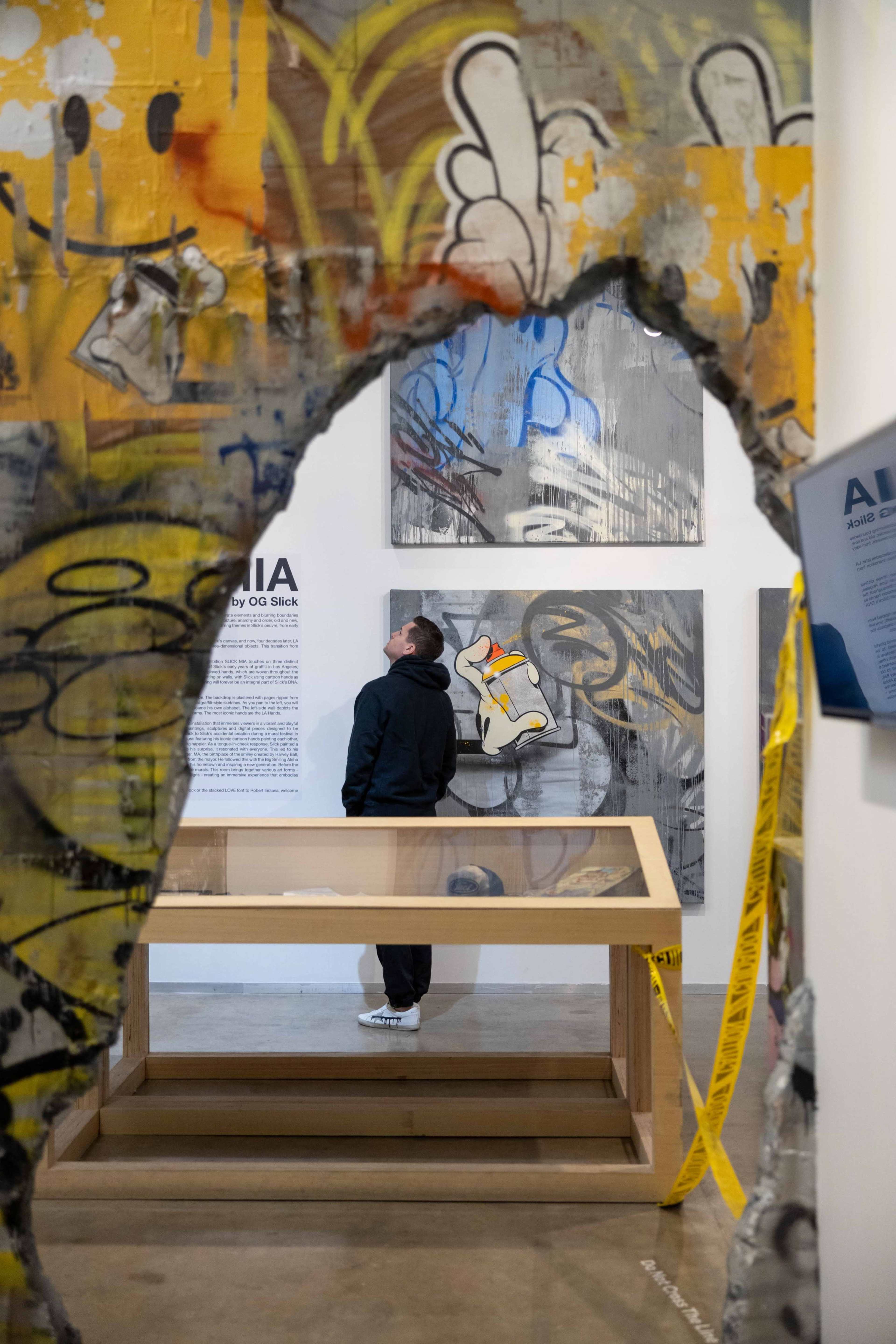 A man stands inside an art gallery, gazing at graffiti artworks displayed on the wall, framed by a hole in a painted structure.