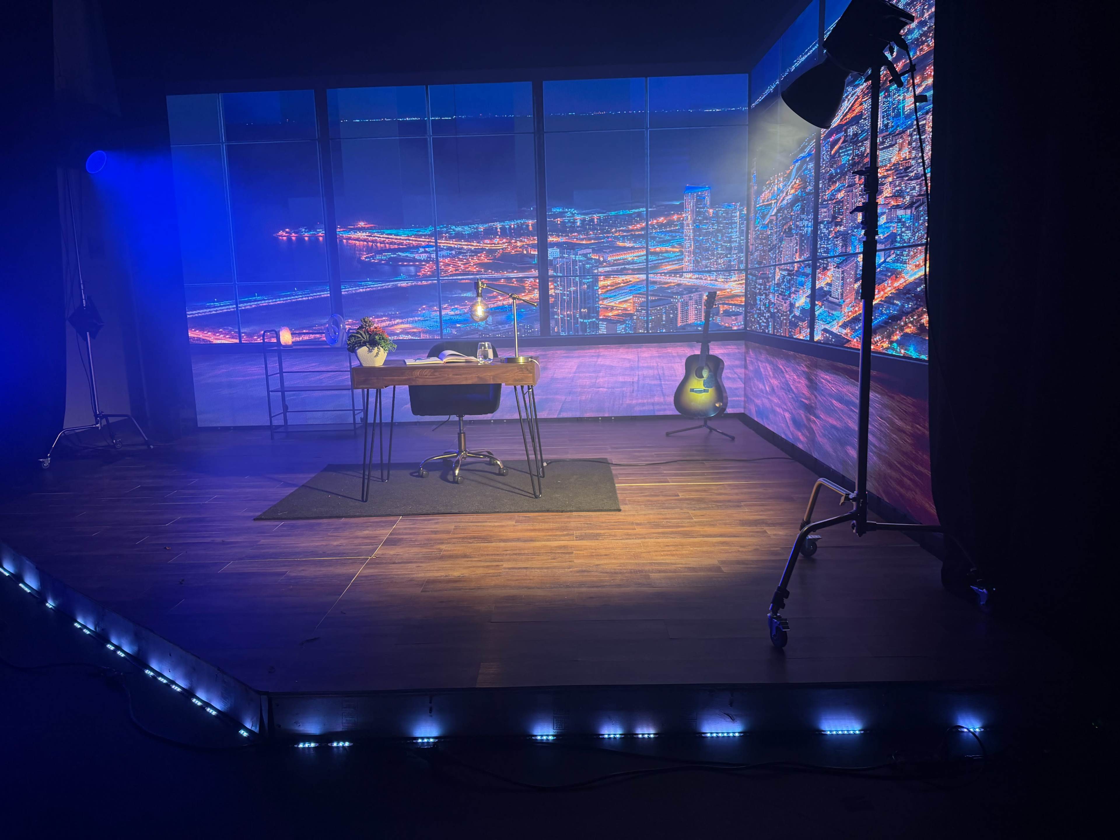 The image depicts a studio set featuring a desk with a chair, a plant, and a guitar, against a backdrop of a brightly lit cityscape.