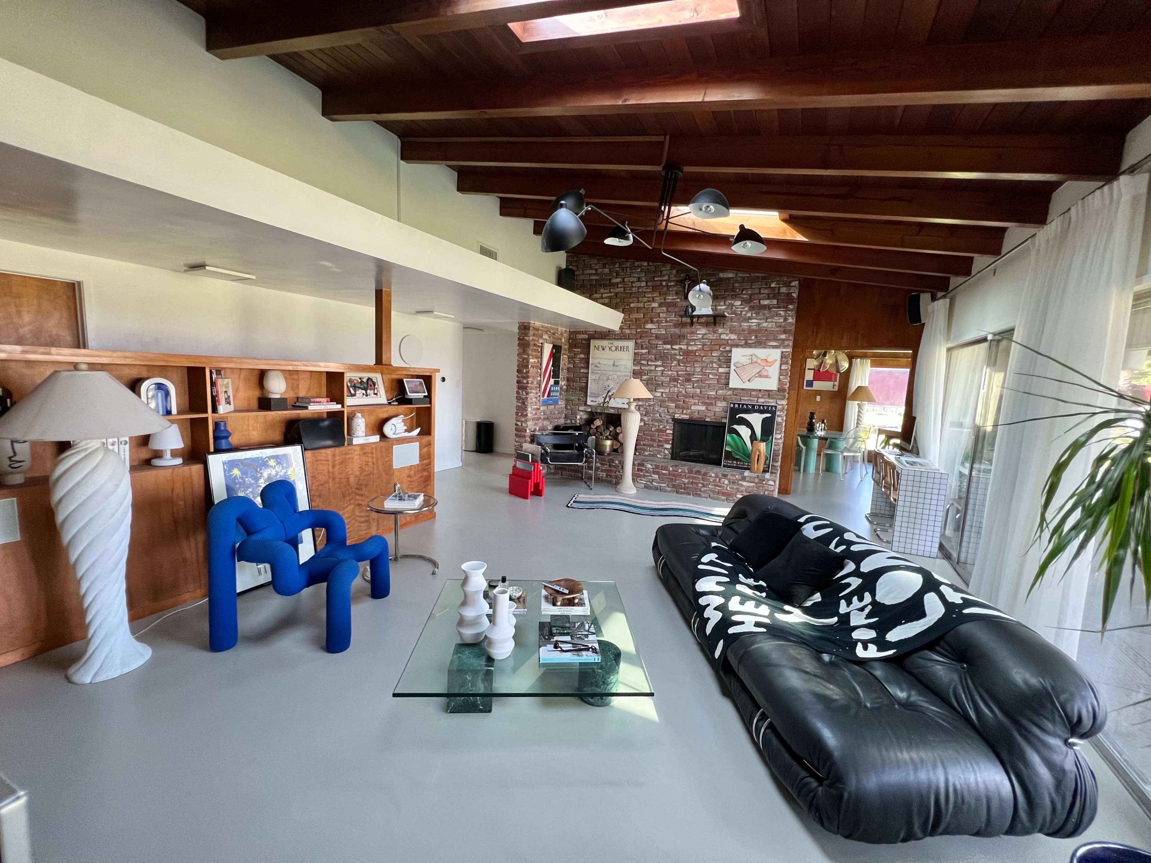 The image shows a spacious living room with a mix of modern furniture, including a blue chair, a black sofa with white patterns, and a glass coffee table, surrounded by wooden beams and a brick wall.