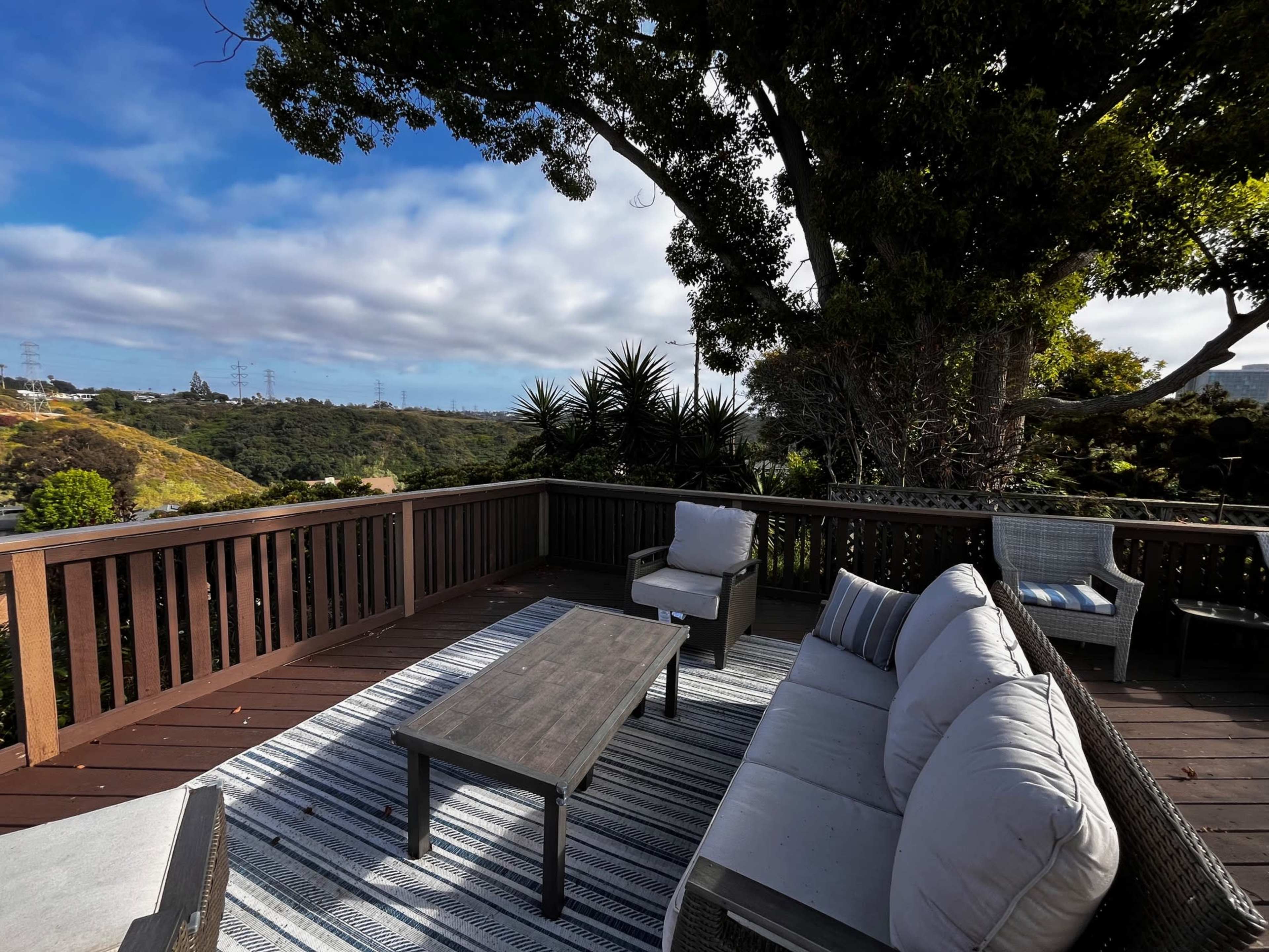 A wooden deck with outdoor furniture, including a sofa and a coffee table, overlooking a hilly landscape under a partly cloudy sky.