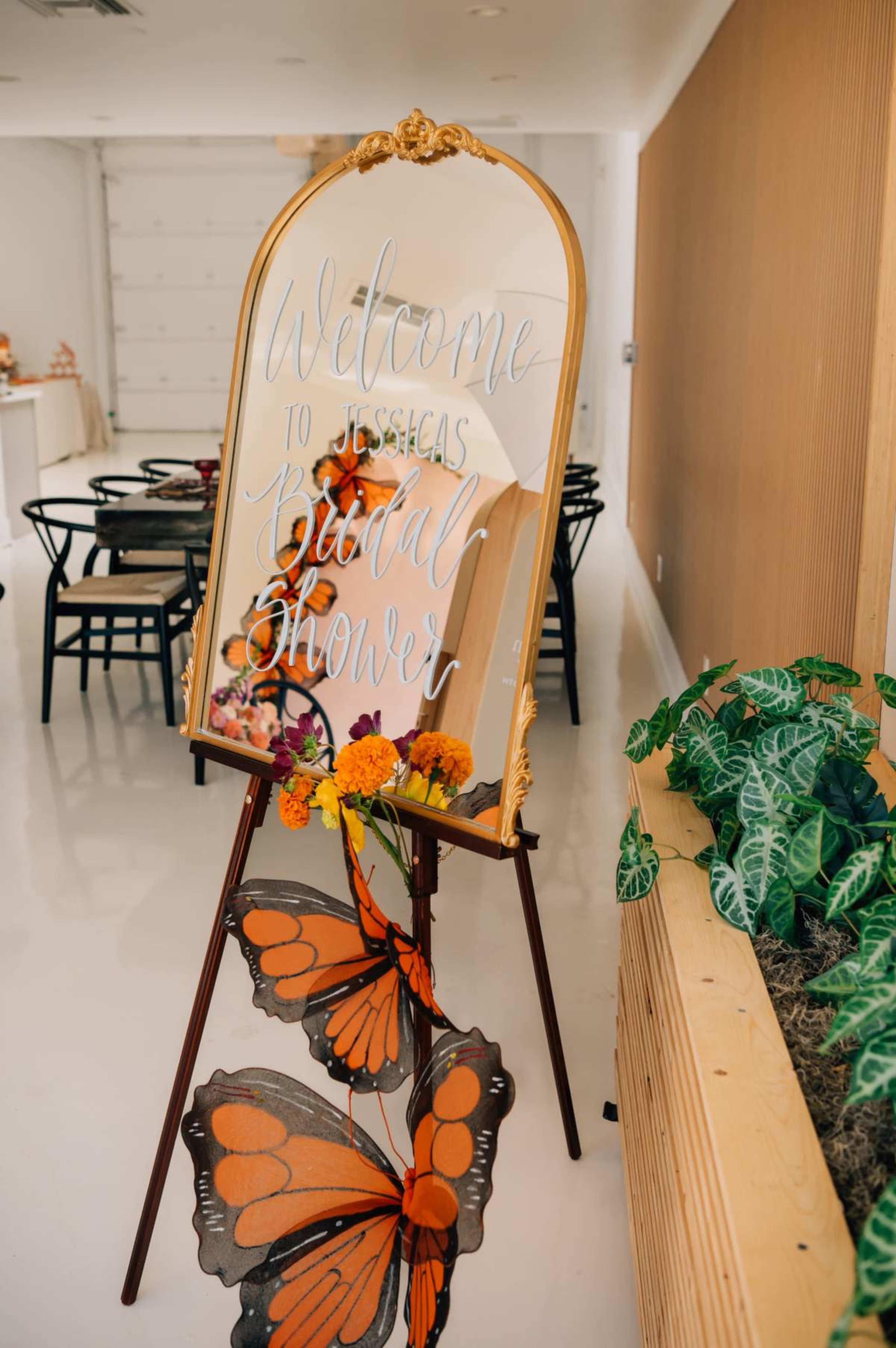 A large ornate mirror displays a welcome sign for a bridal shower, surrounded by decorative flowers and a butterfly motif in a well-lit indoor space.