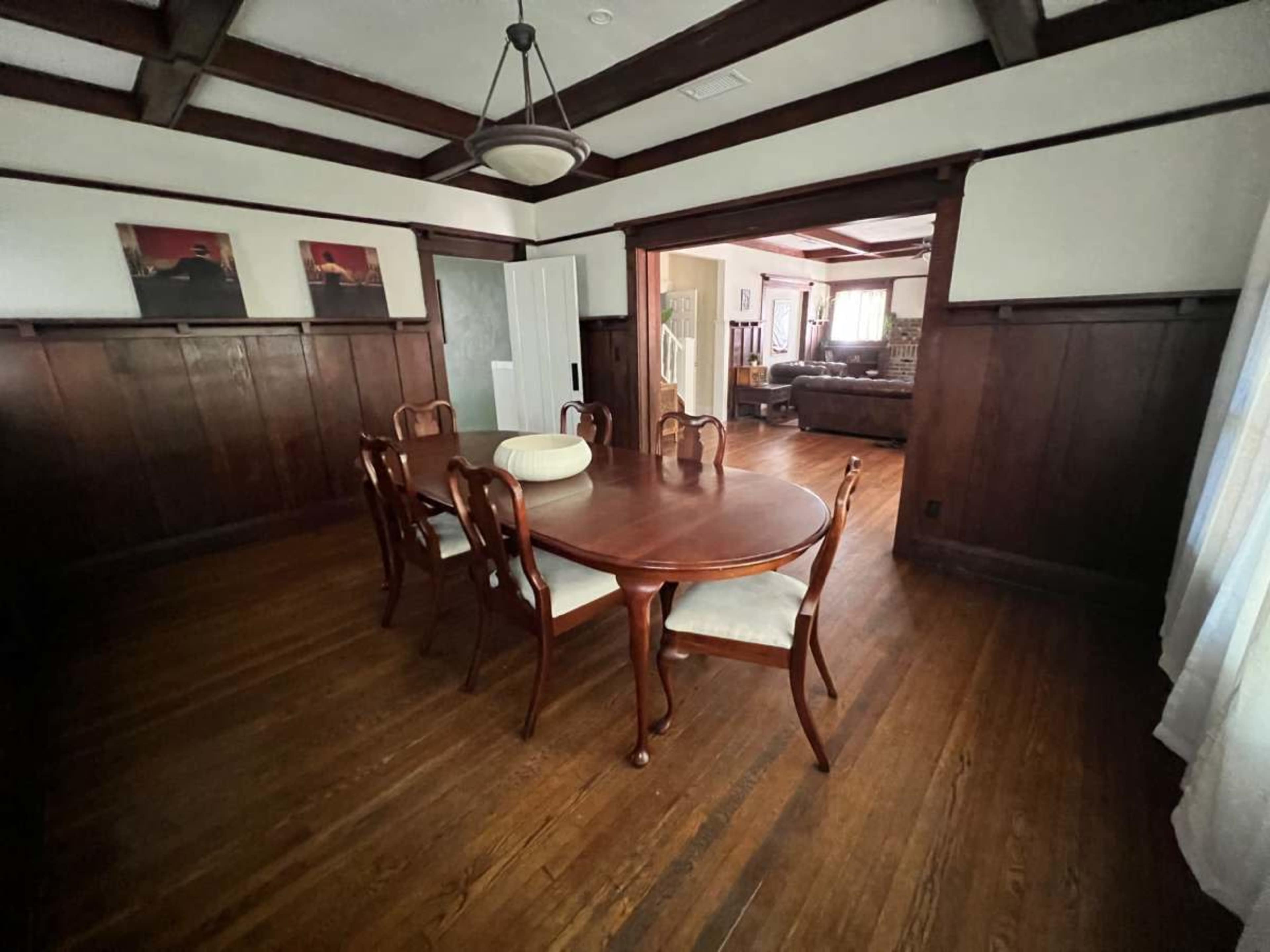 The image shows a dining room with a wooden table and chairs, featuring dark wooden beams and walls, and an entrance leading to another room.