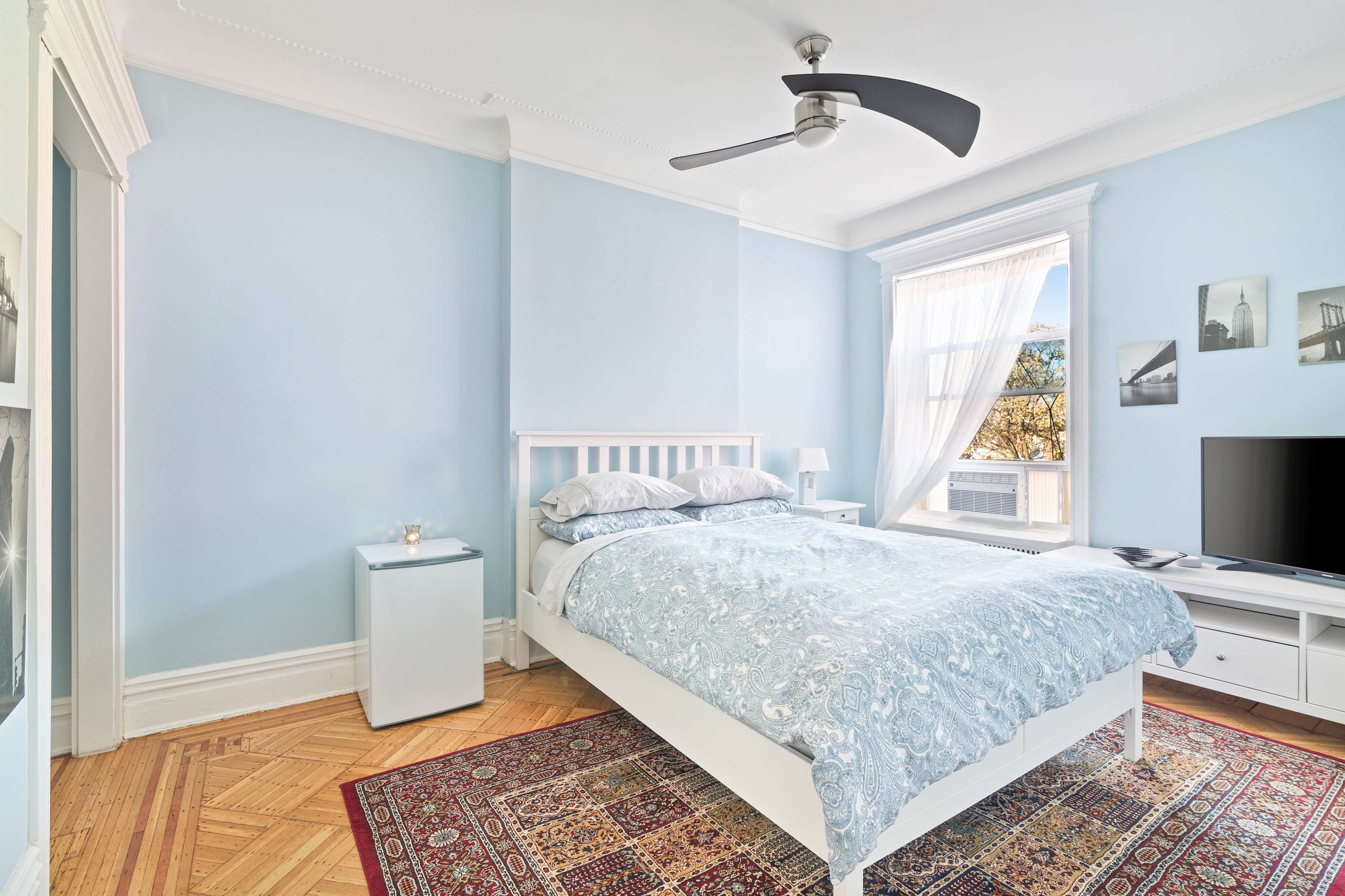 A light blue bedroom features a white bed with a patterned comforter, a nightstand, a television, and a large window with sheer curtains.