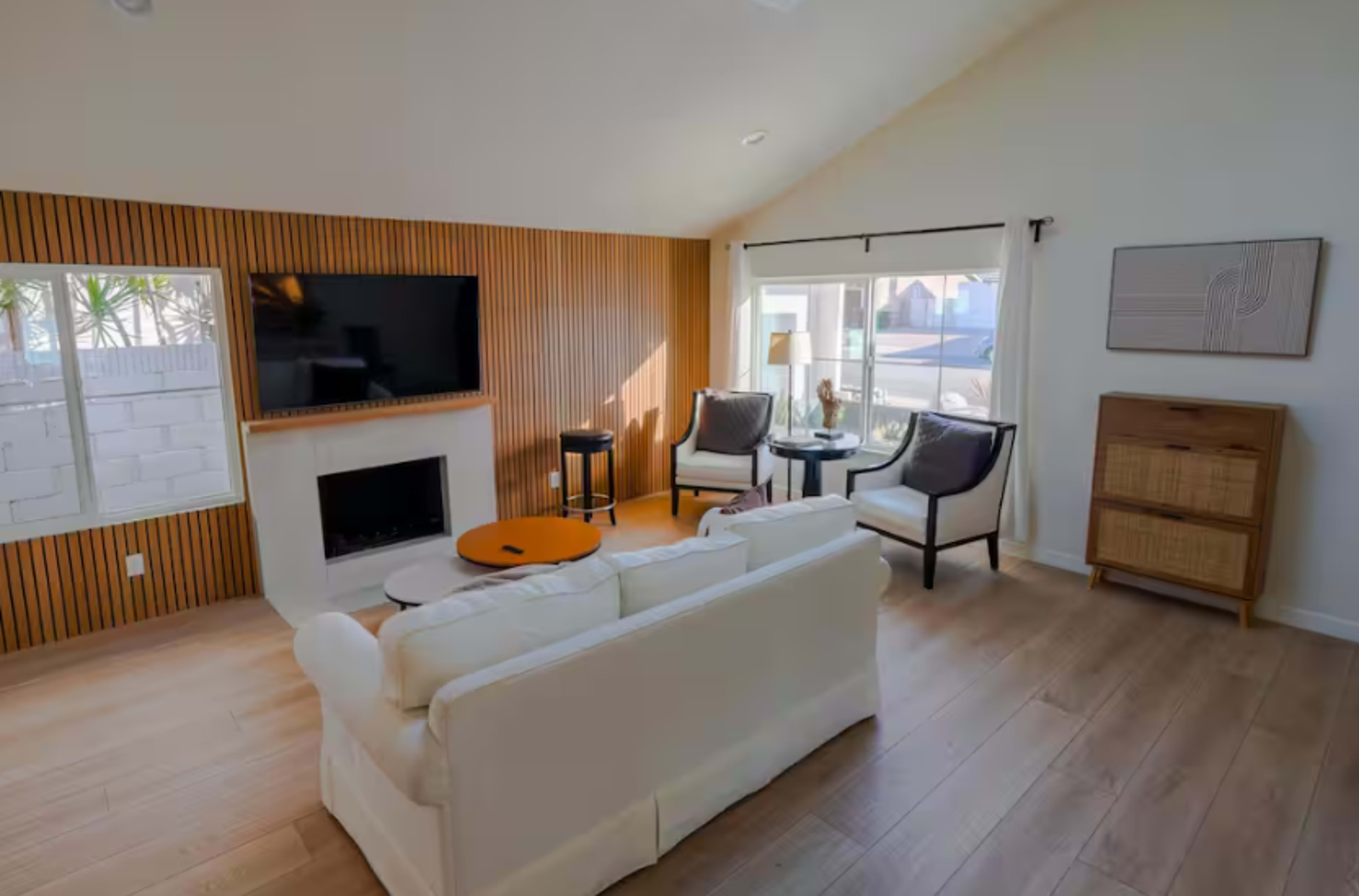 A modern living room features a white sofa, two armchairs, a coffee table, a wall-mounted TV, and wooden paneling.