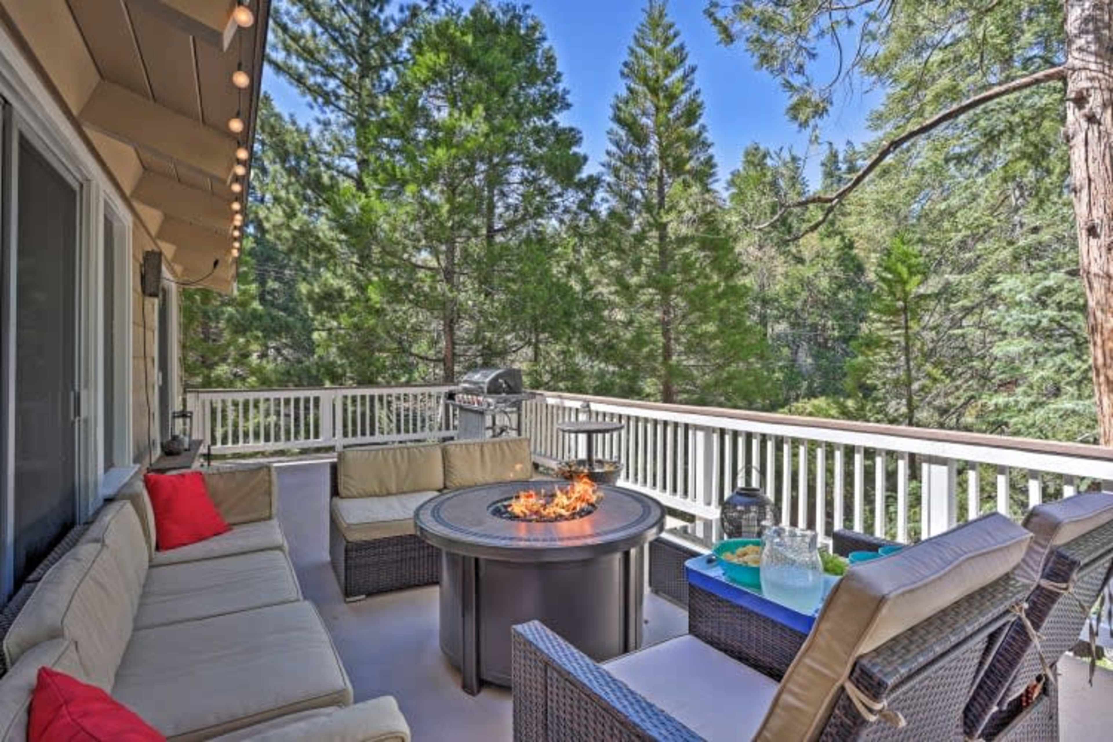 The image shows a deck with outdoor seating, a fire pit, and a backdrop of tall trees.