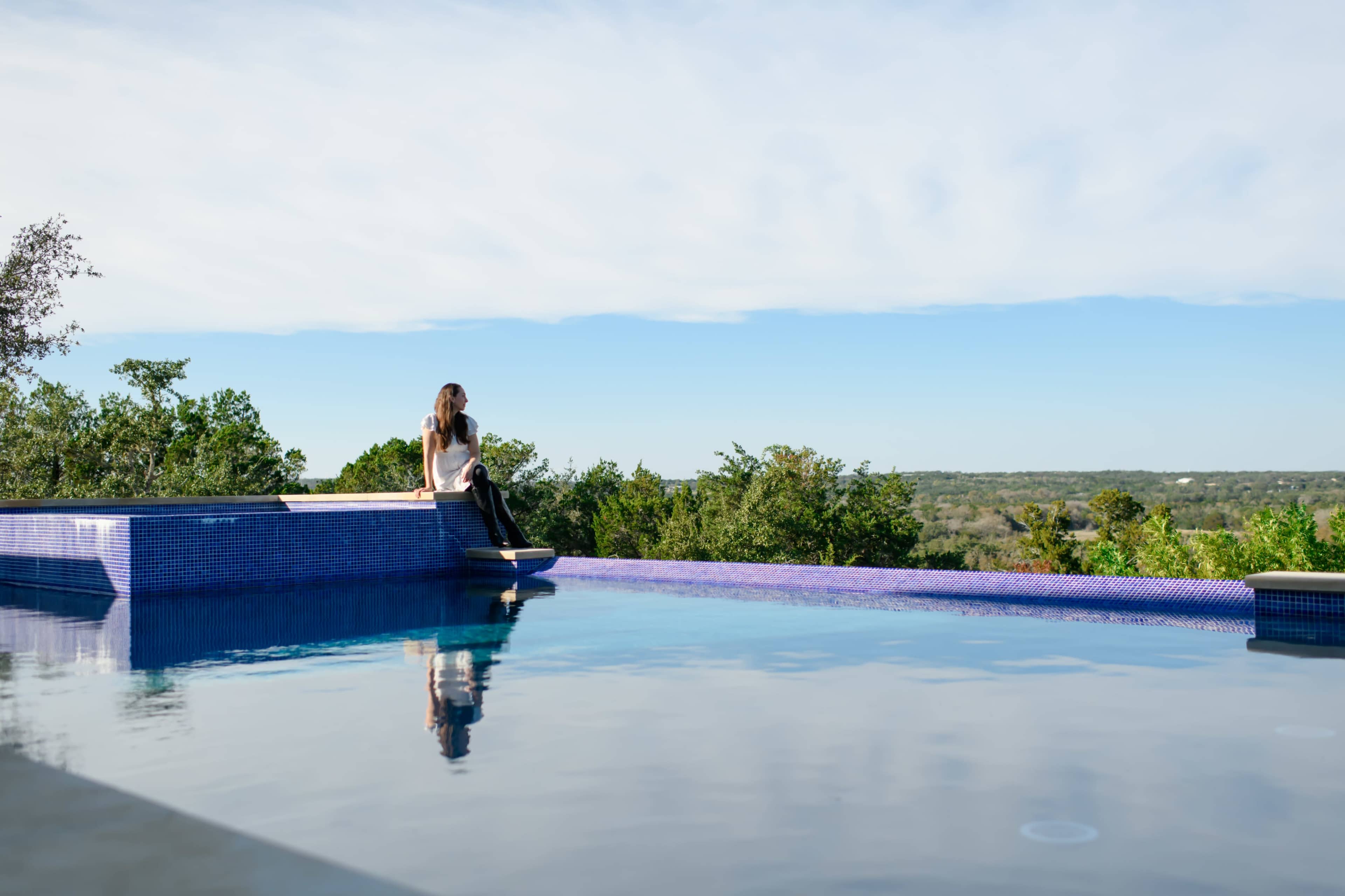 A person sits on the edge of an infinity pool, overlooking a green landscape under a clear blue sky.