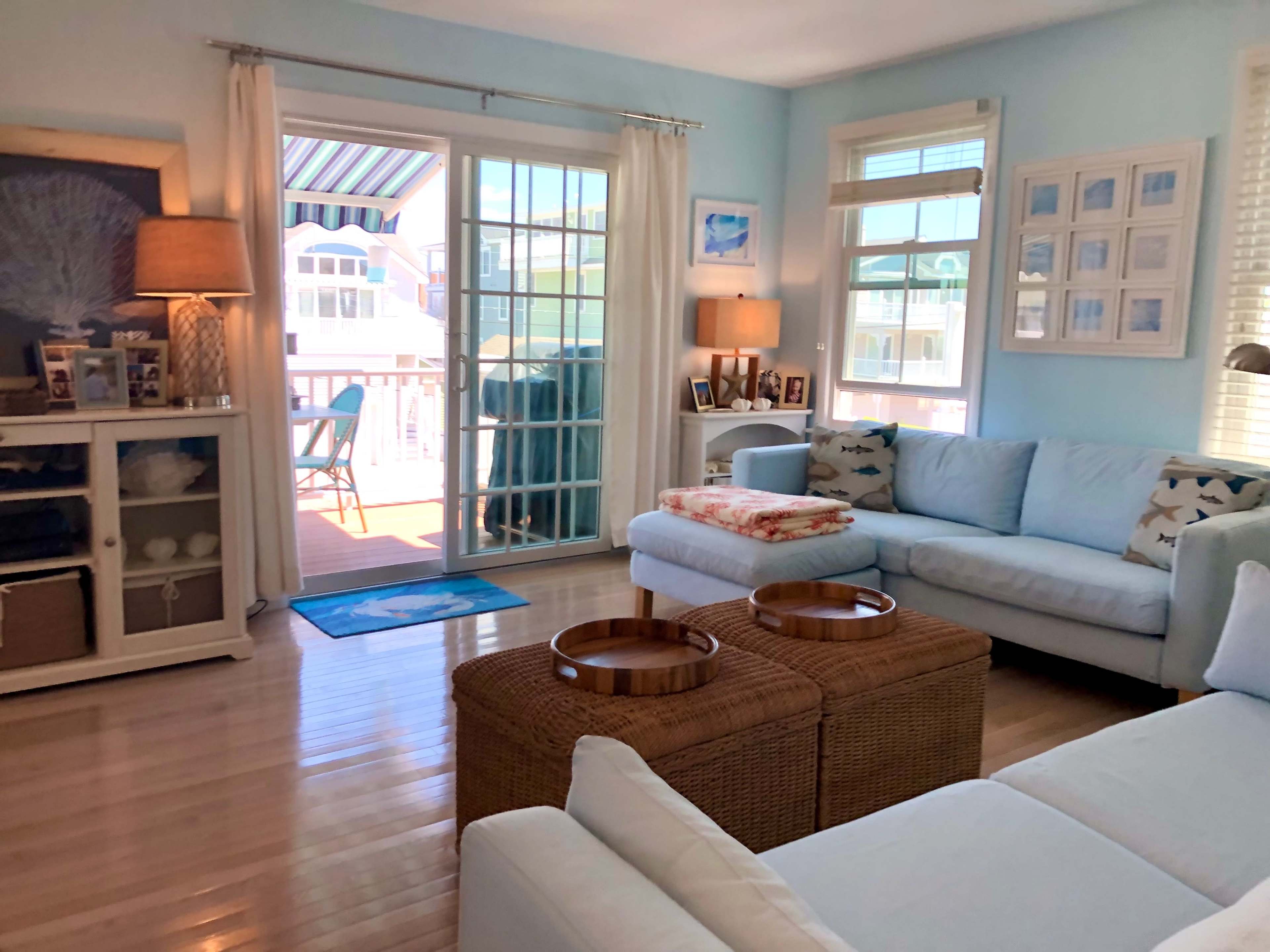 A bright living room features a light blue color scheme, a comfortable sectional couch, two woven baskets as coffee tables, and a sliding glass door opening onto a deck with outdoor seating.