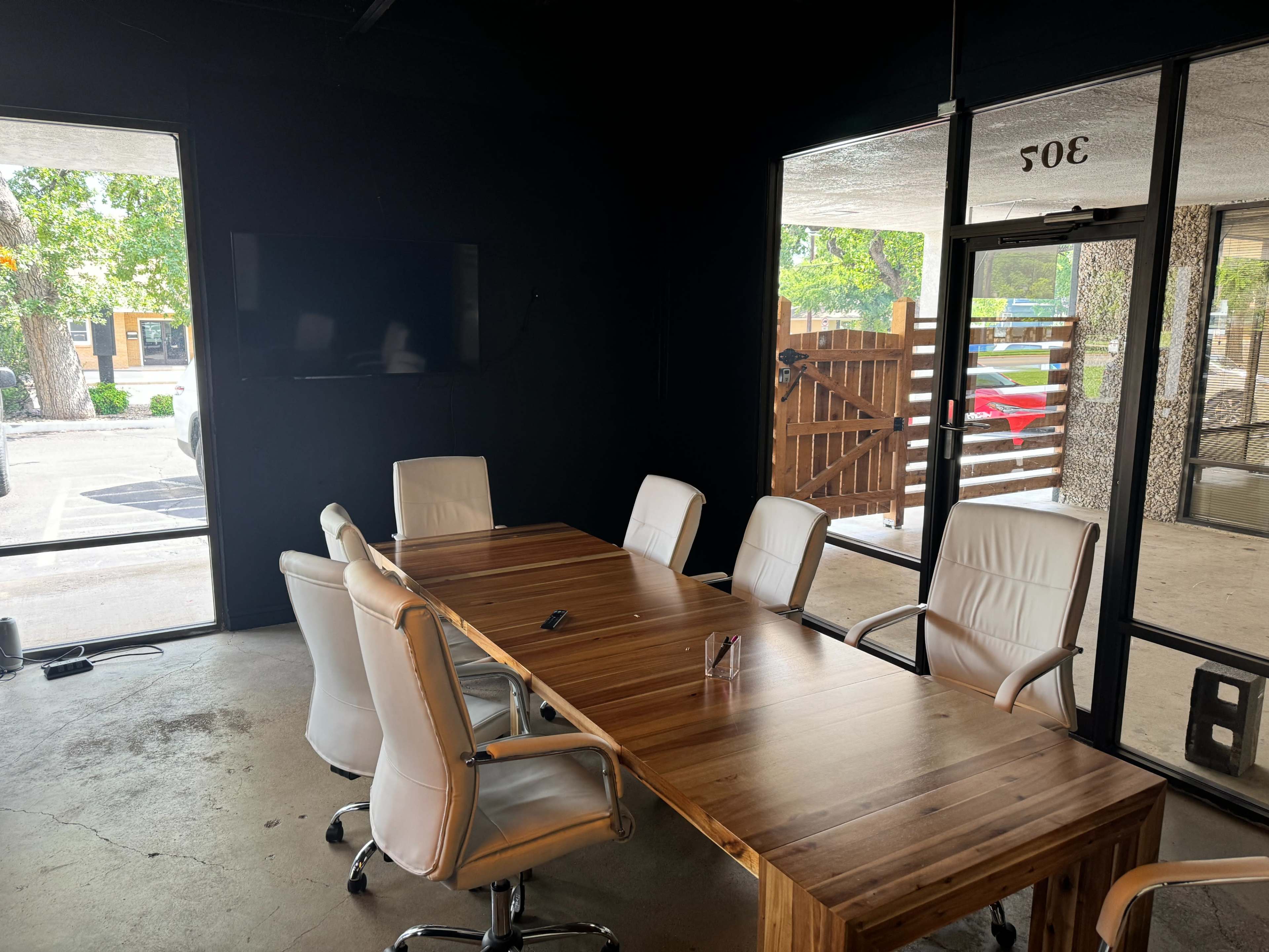 A conference room features a wooden table surrounded by white chairs, with large glass windows letting in natural light.