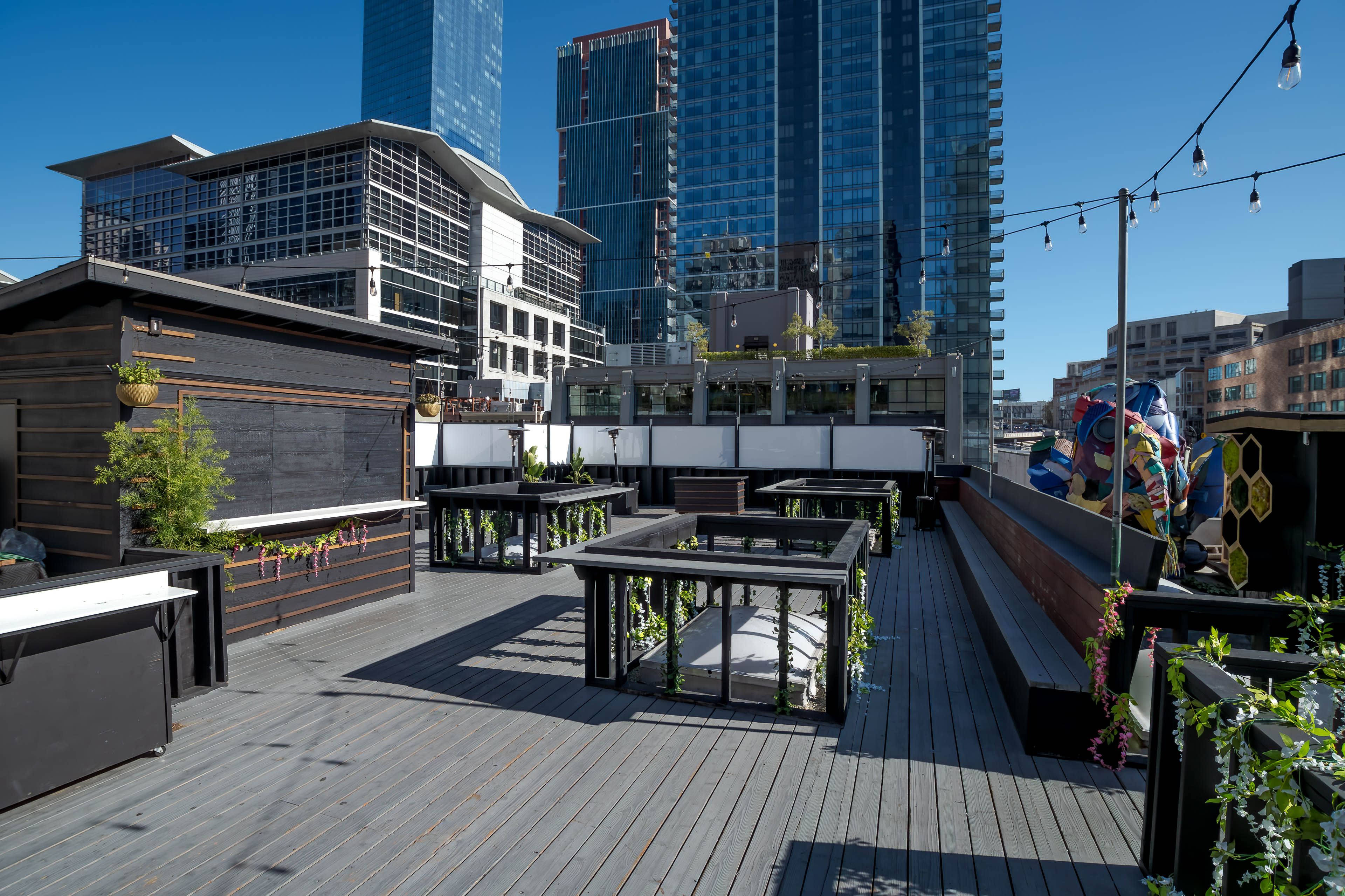 A rooftop area with wooden decking, garden planters, and structures set against a backdrop of modern high-rise buildings under a clear blue sky.