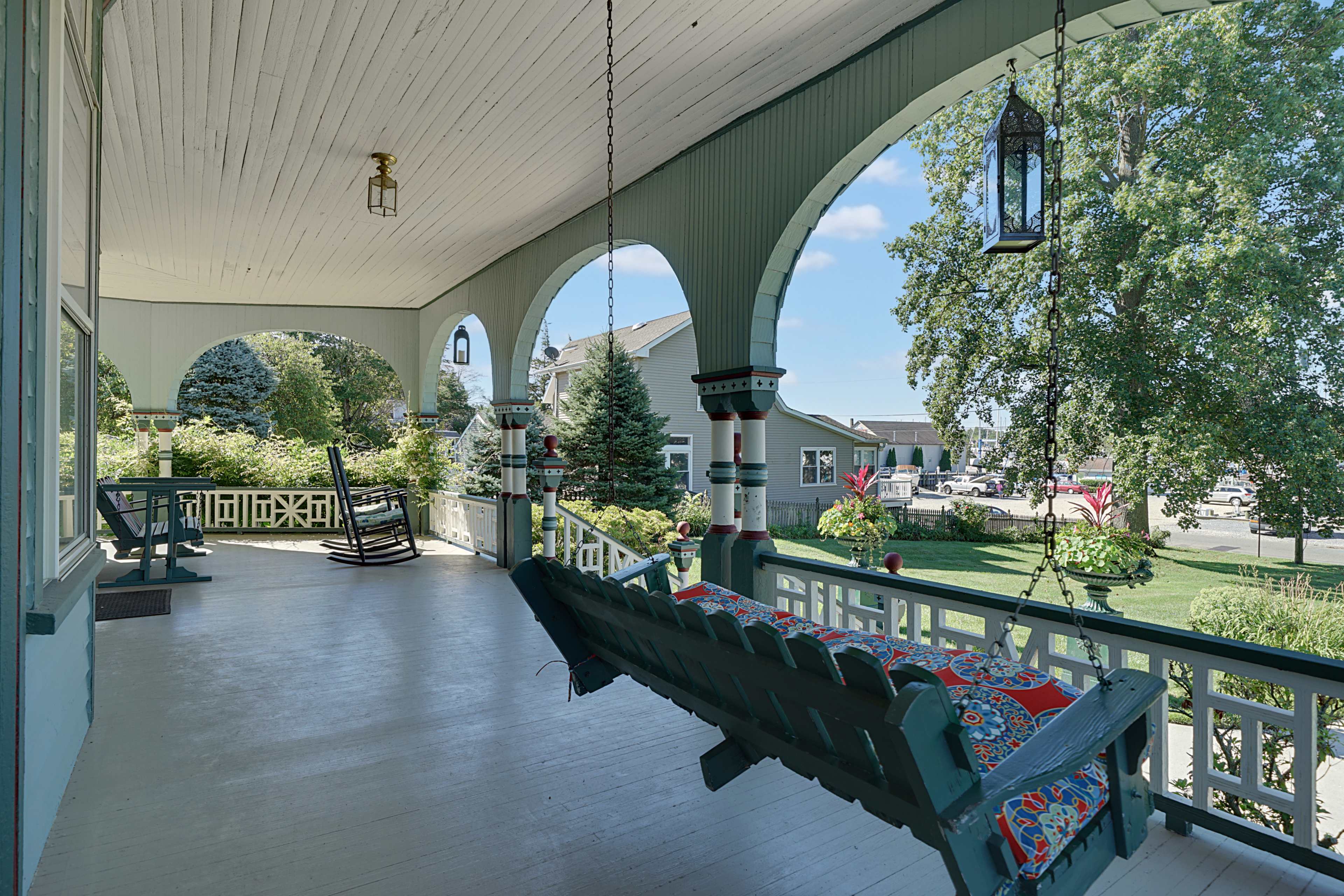 A covered porch with a swing, rocking chairs, and decorative lanterns, framed by arches and surrounded by greenery.