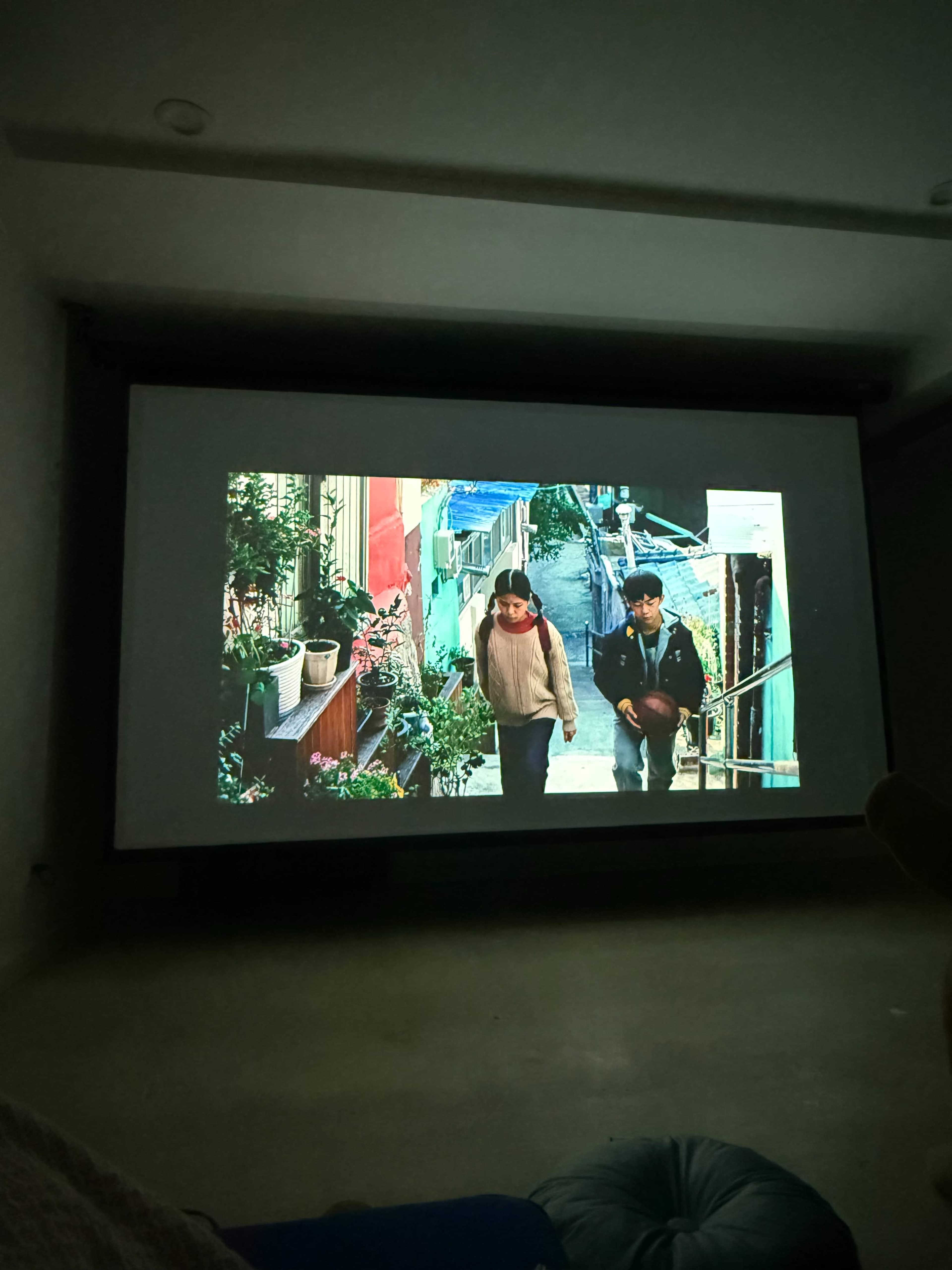 The image shows a scene from a movie projected on a large screen, featuring two characters walking down a colorful street lined with plants and buildings.