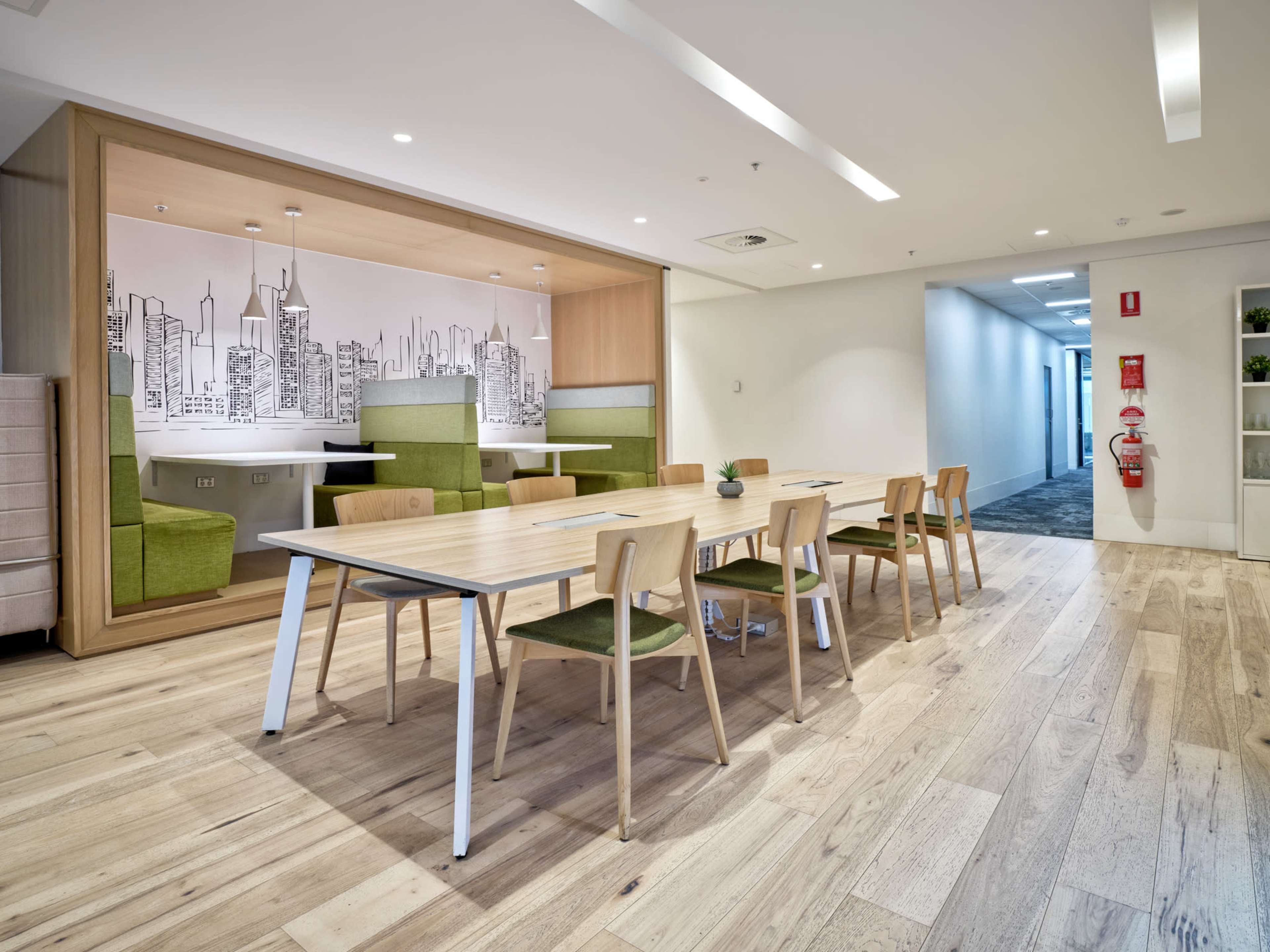The image shows a modern office space featuring a wooden table surrounded by several chairs, with a wall mural of a city skyline in the background and a seating nook to the side.