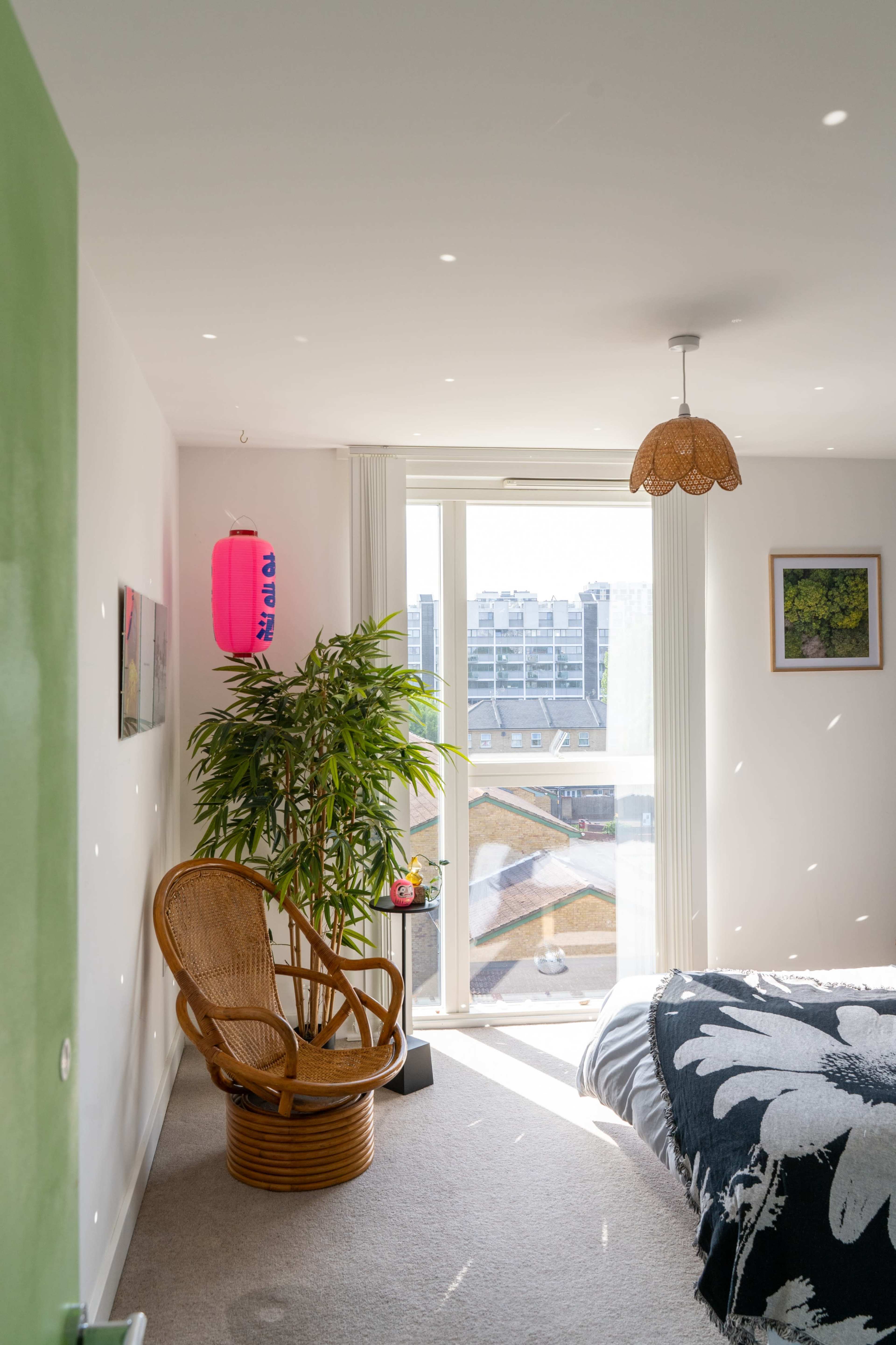 A cozy bedroom features a rattan chair beside a large window with city views, a small plant, and decorative elements.