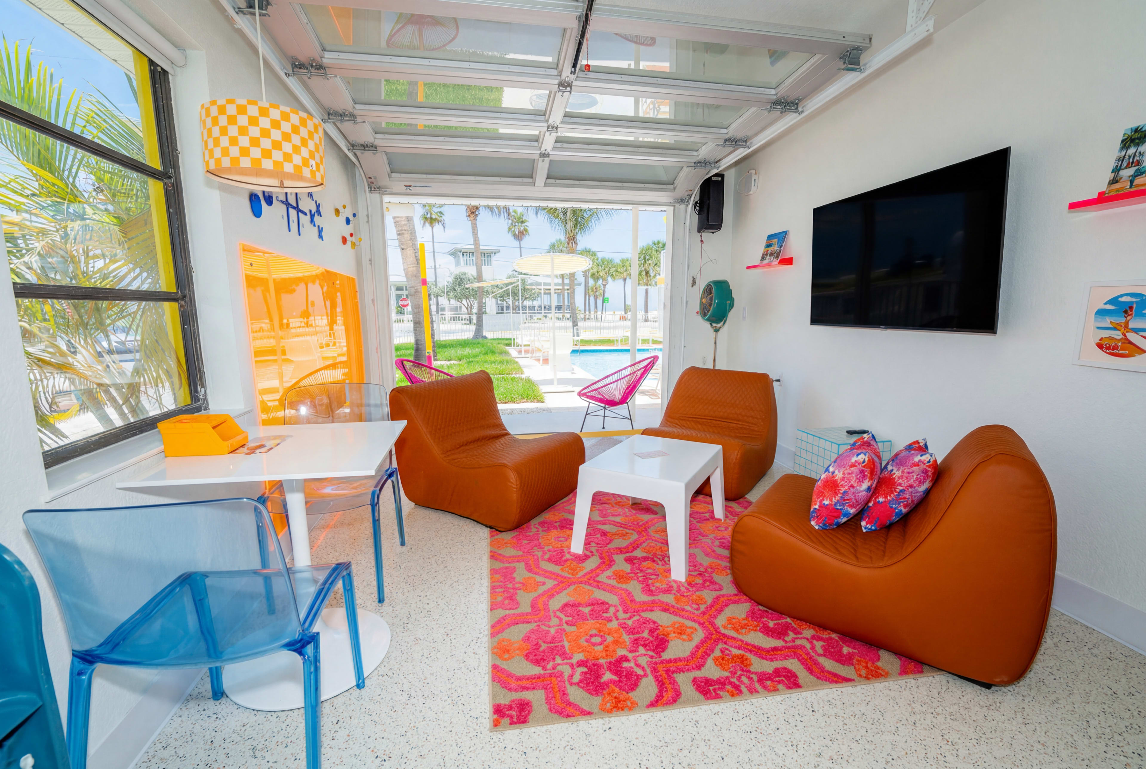 The image shows a modern indoor lounge area with colorful furniture, a large television mounted on the wall, and a view of a pool outside through large garage-style windows.