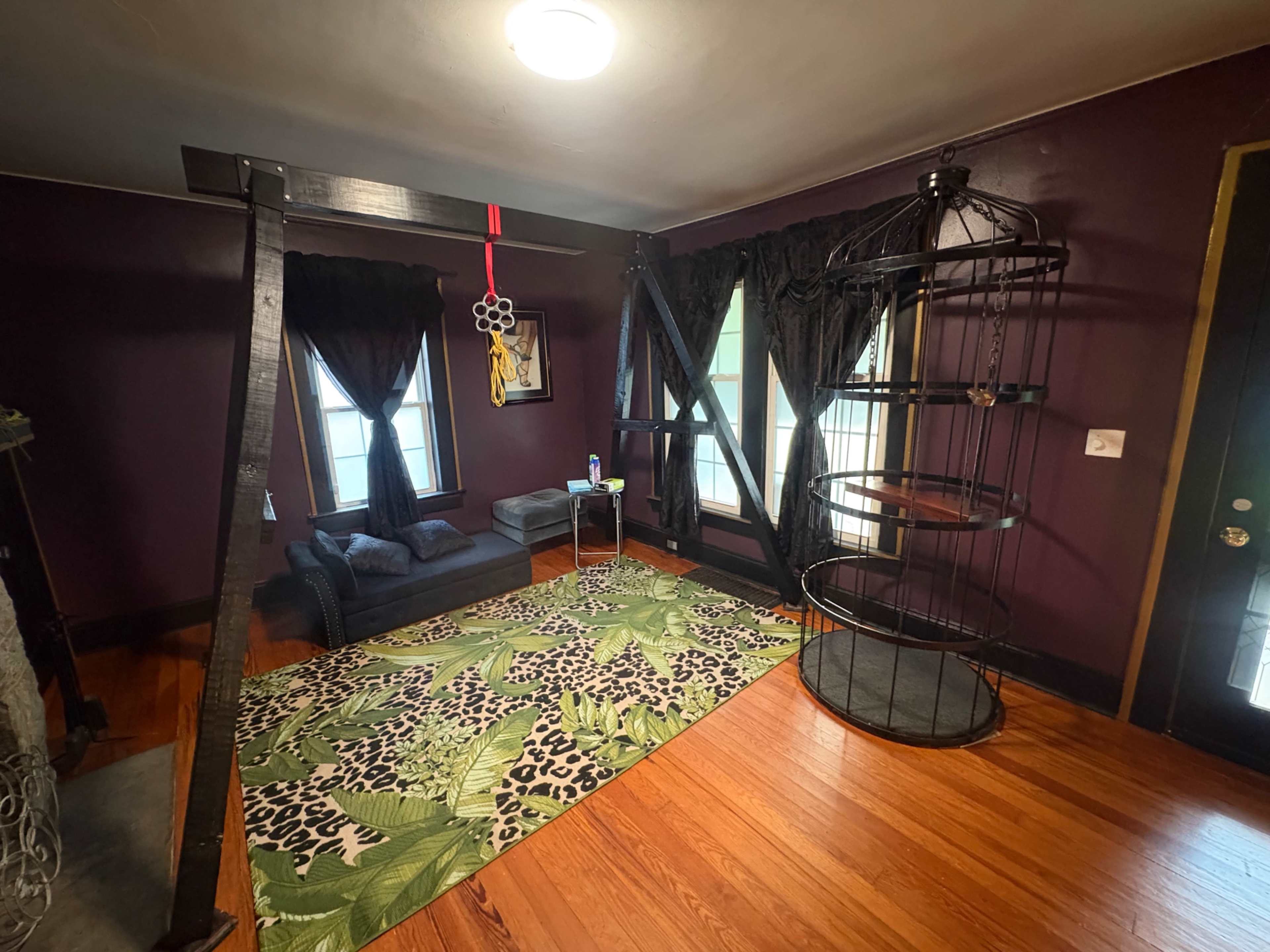 The room features dark purple walls, a patterned rug, a modern seating area with a small table, and a unique, circular shelf structure beside large windows dressed with black curtains.