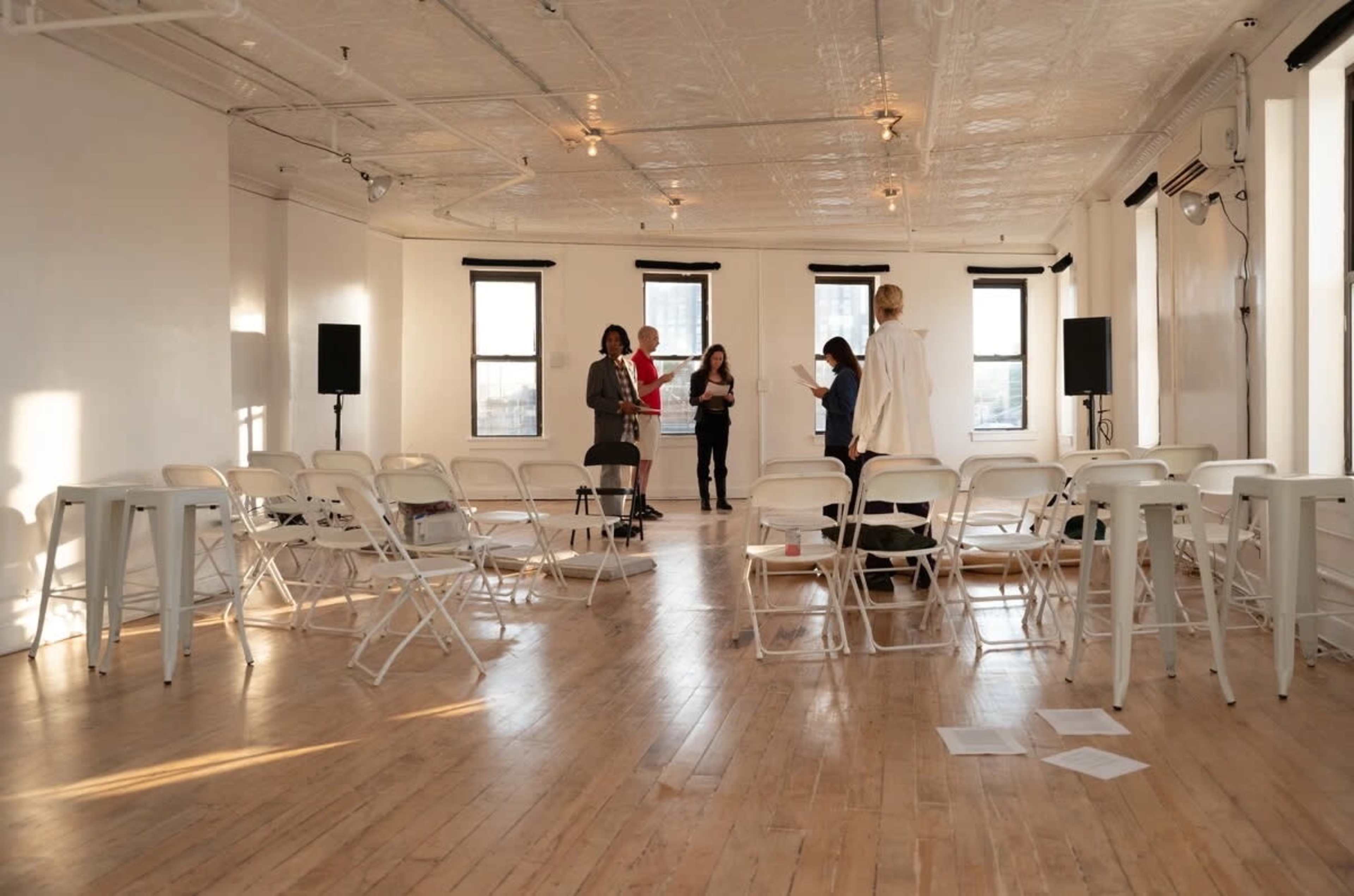 A spacious room with wooden flooring is set up with rows of white folding chairs, where four people are gathered near the center.