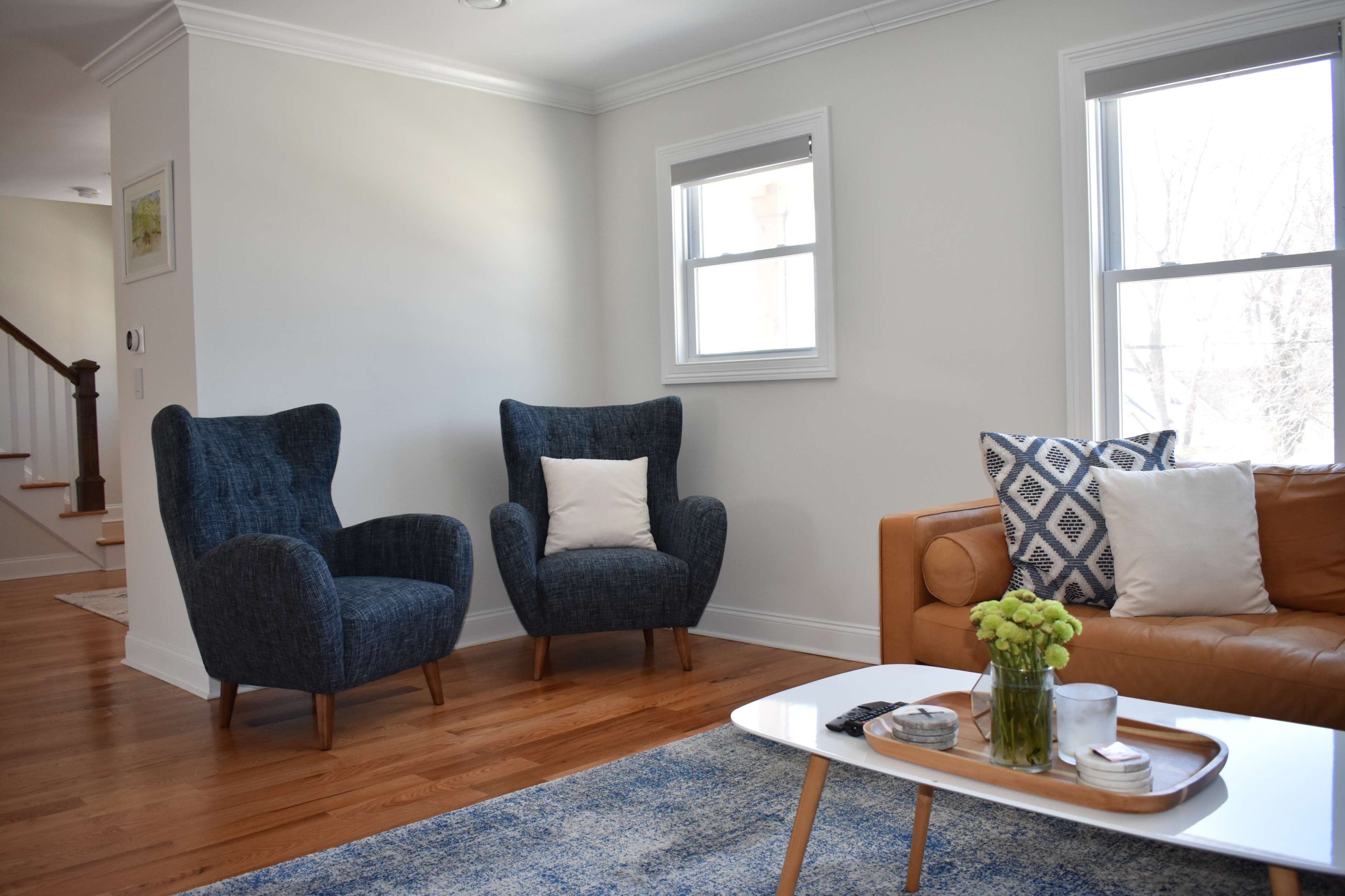 The image shows a bright, modern living room featuring two blue armchairs and a brown leather sofa, with a coffee table and a rug on a wooden floor.