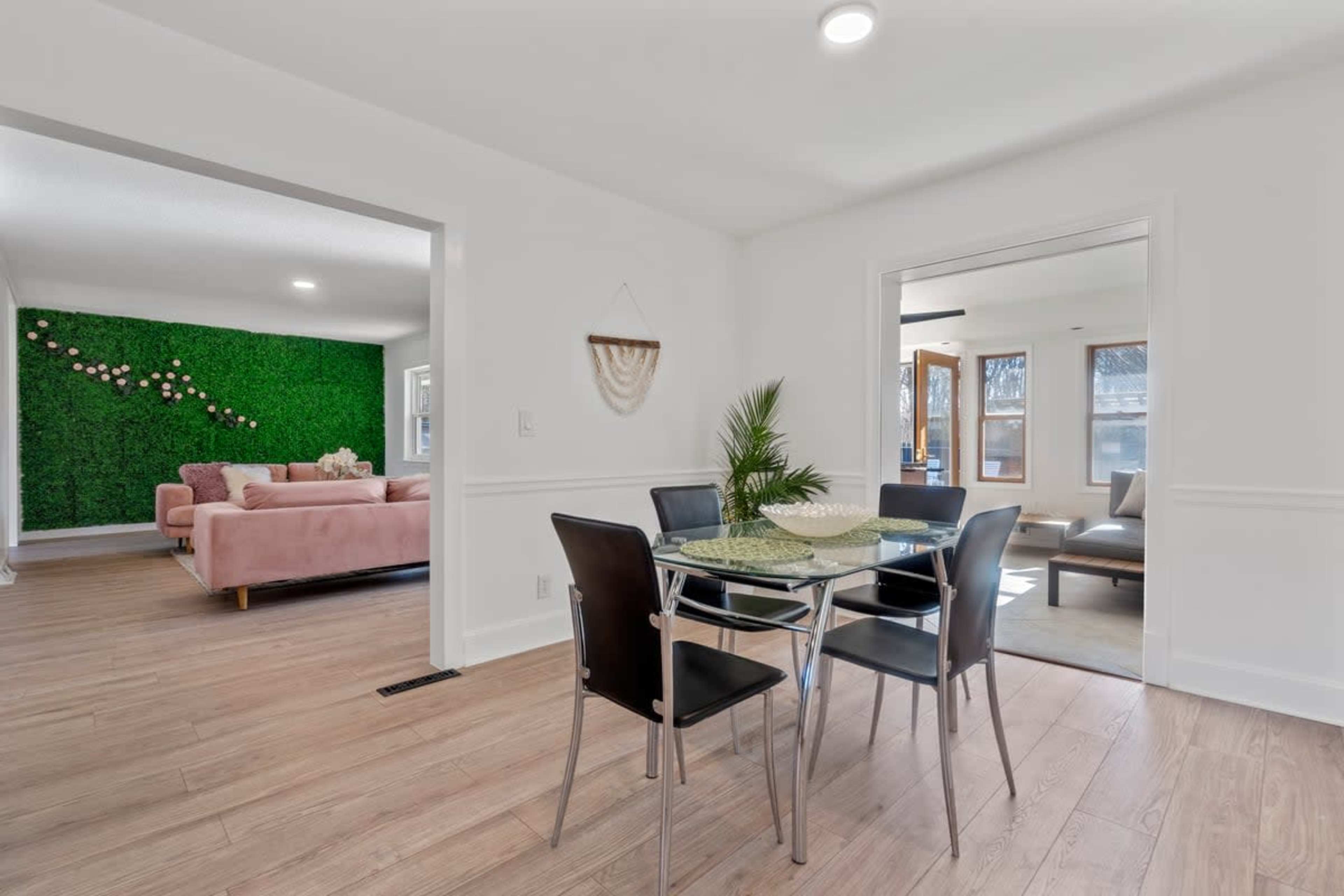 The dining area features a glass table surrounded by black chairs, with a view of a living room that has green plant wall decor and a pink sofa.