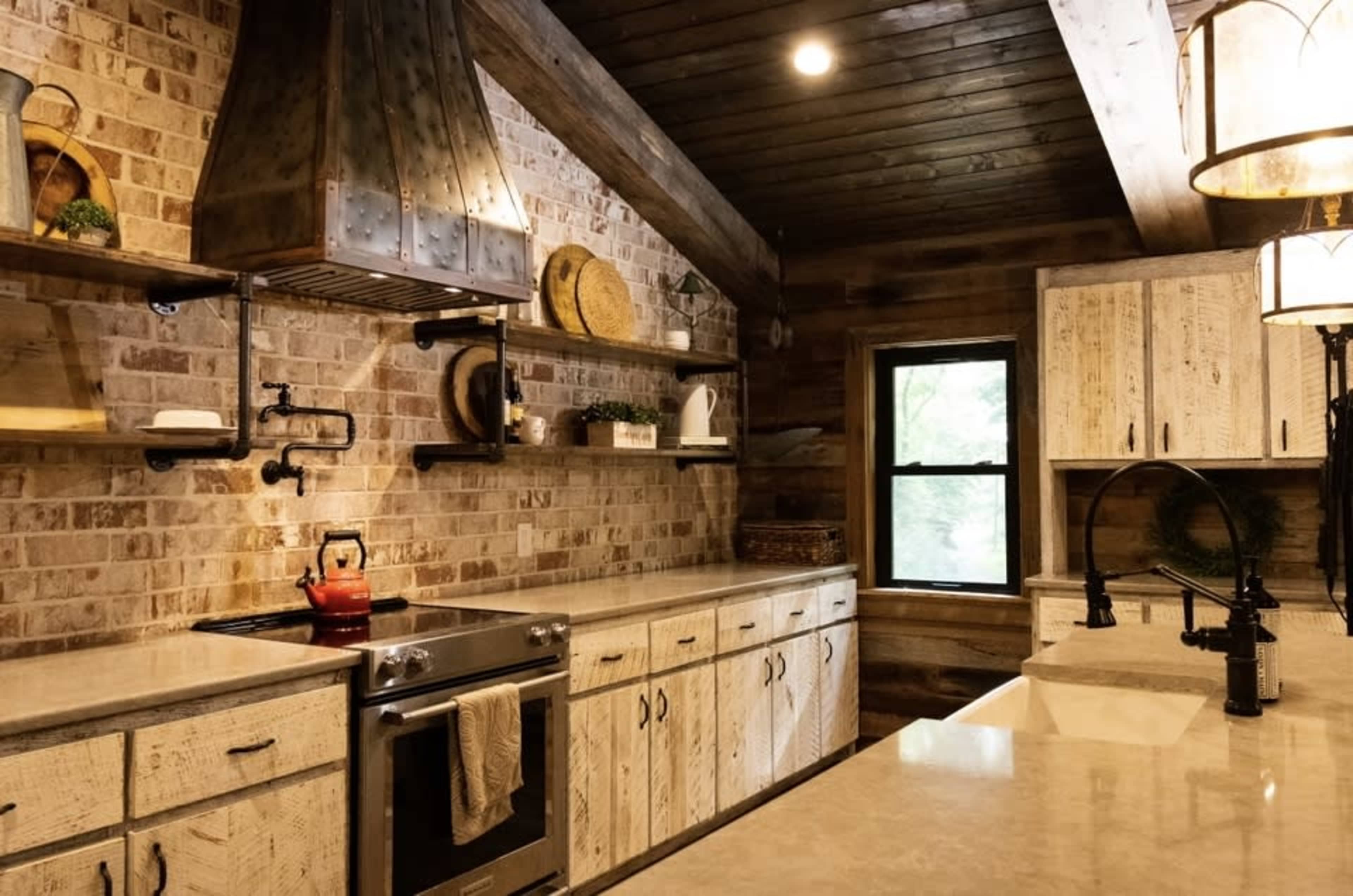A rustic kitchen with wooden cabinetry, a stainless steel stove, a brick wall, and overhead shelves displaying decorative items.