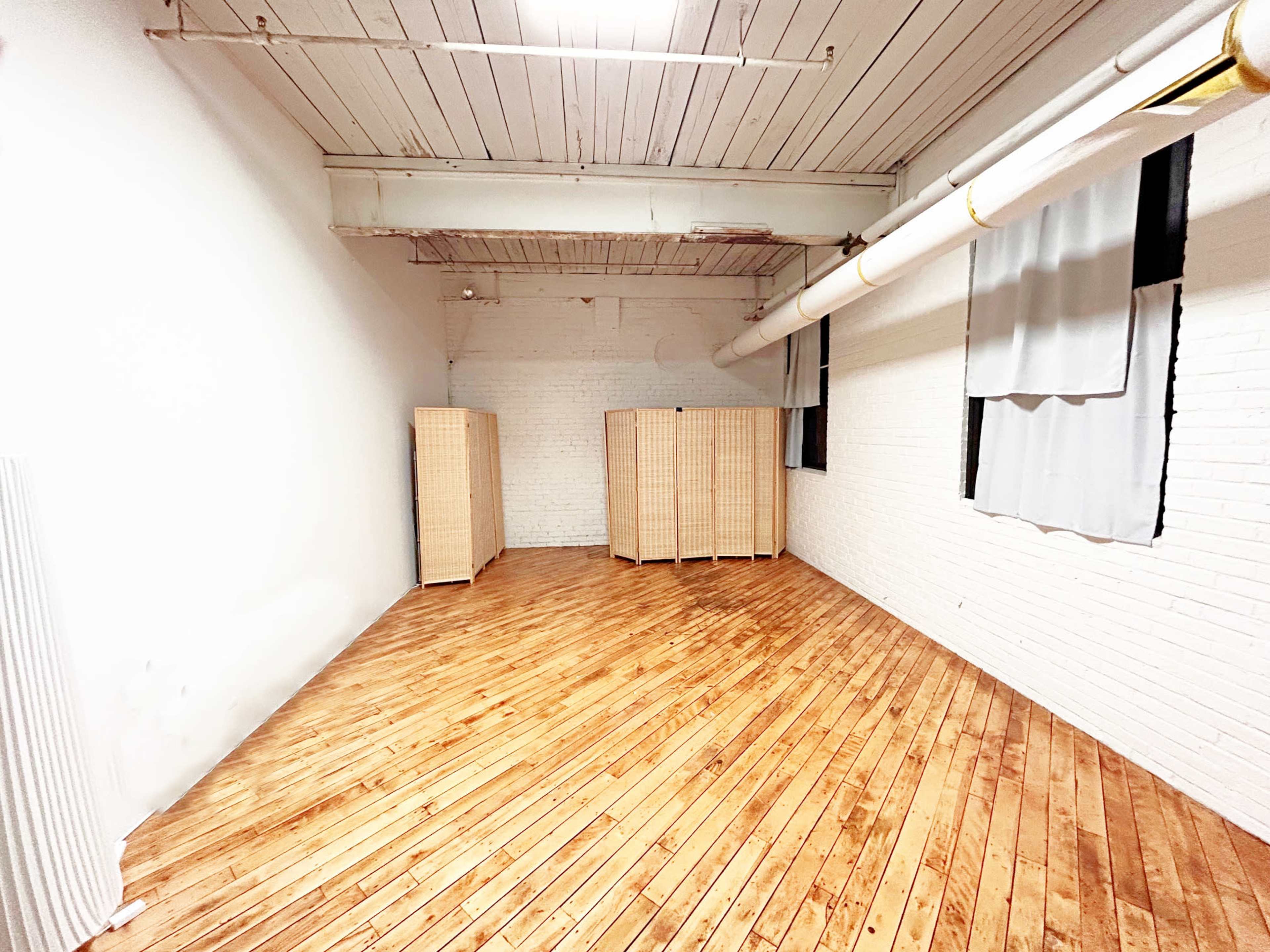 The image shows a spacious, empty room with wooden flooring, white brick walls, and three folding screens positioned in the center.