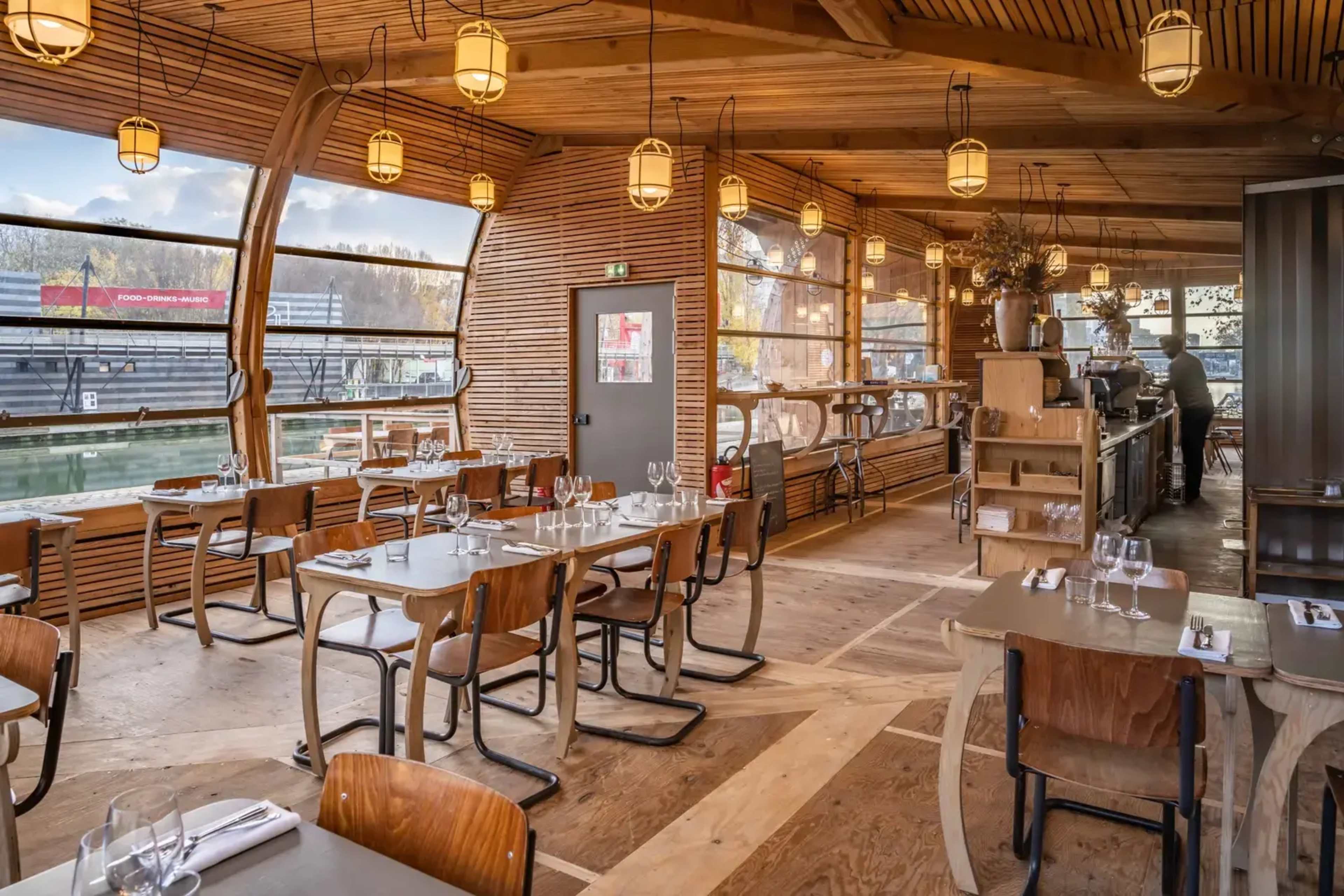 A modern restaurant with wooden interiors, featuring tables and chairs arranged near large windows overlooking a waterfront.