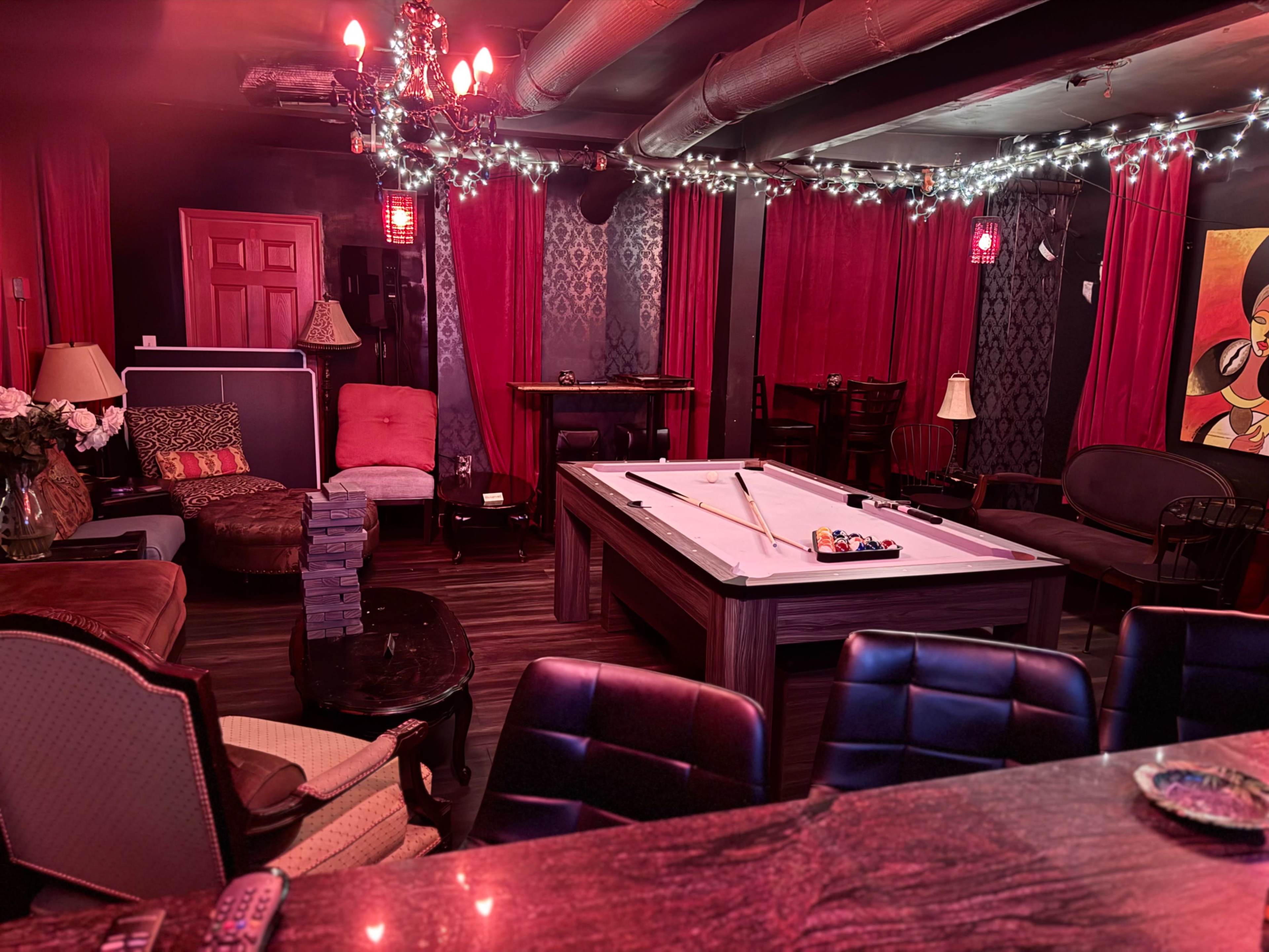 The image shows a cozy lounge area featuring a pool table, various seating arrangements, and red curtains with decorative lighting.