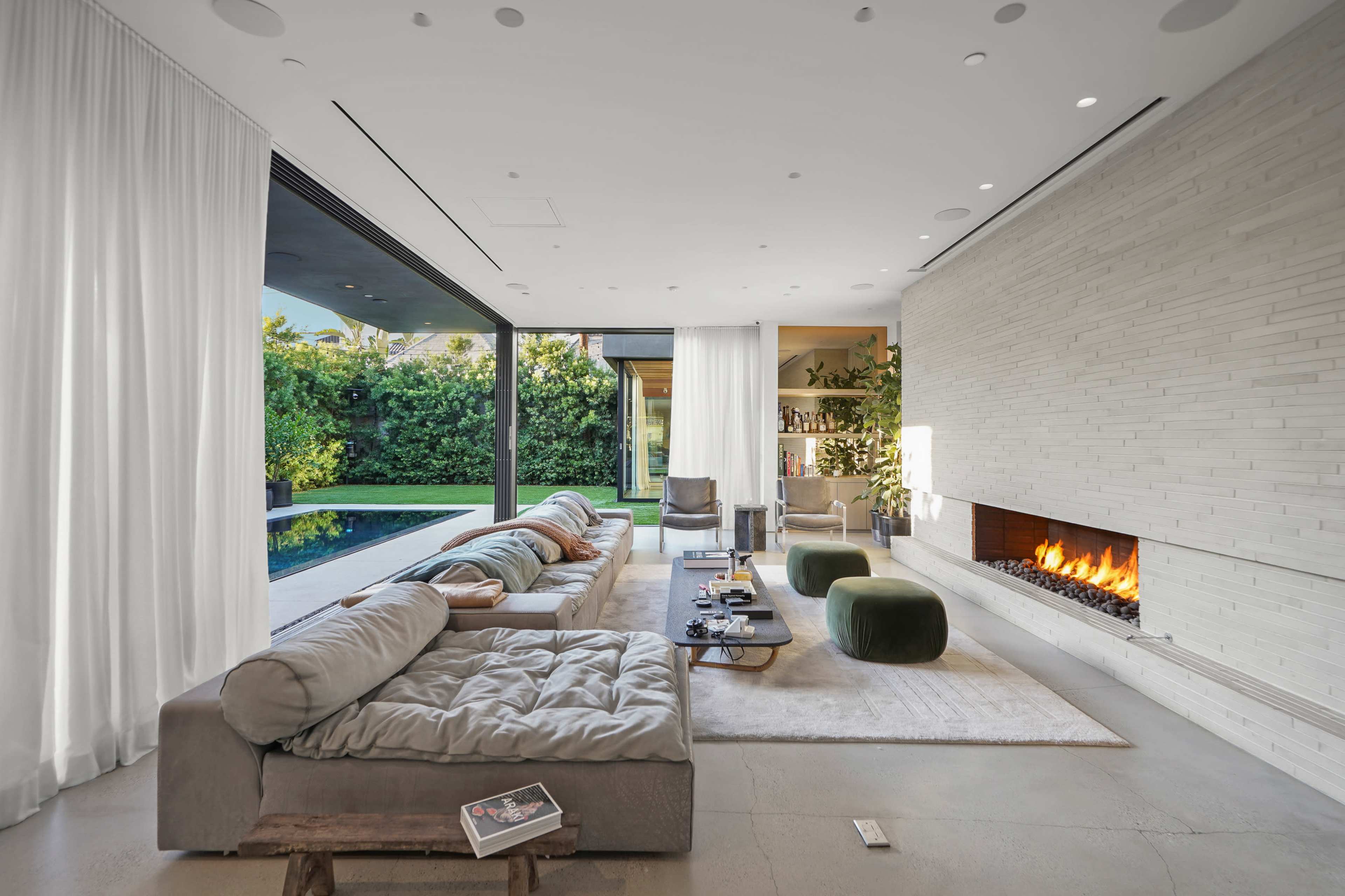 The image depicts a modern living room featuring a large sectional sofa, a minimalistic fireplace, and floor-to-ceiling windows that open to a view of greenery outside.