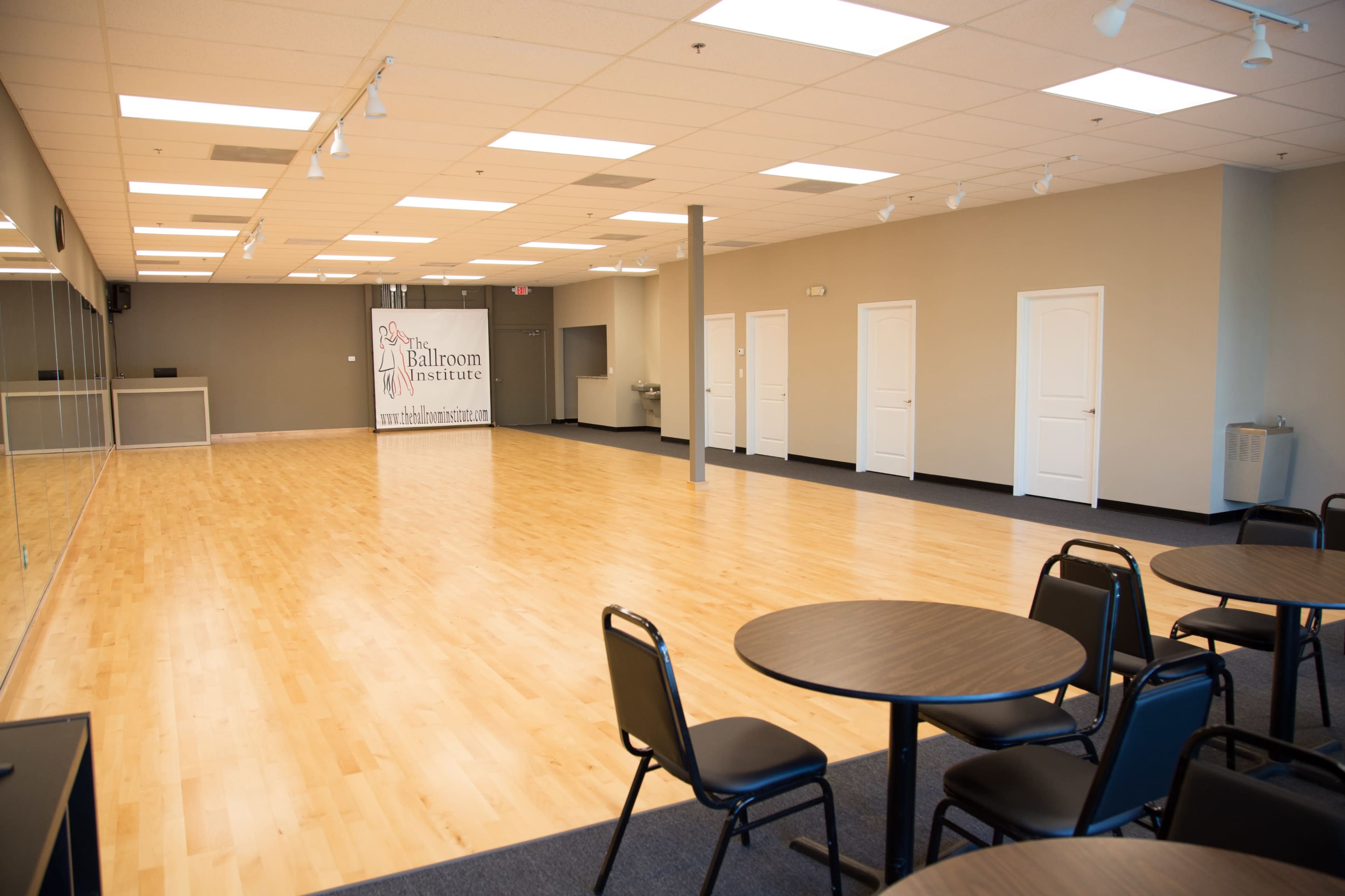 The image shows a spacious dance studio with wooden flooring, large mirrors on one wall, and a sign for a ballroom institute in the background, along with several round tables and chairs arranged in the foreground.