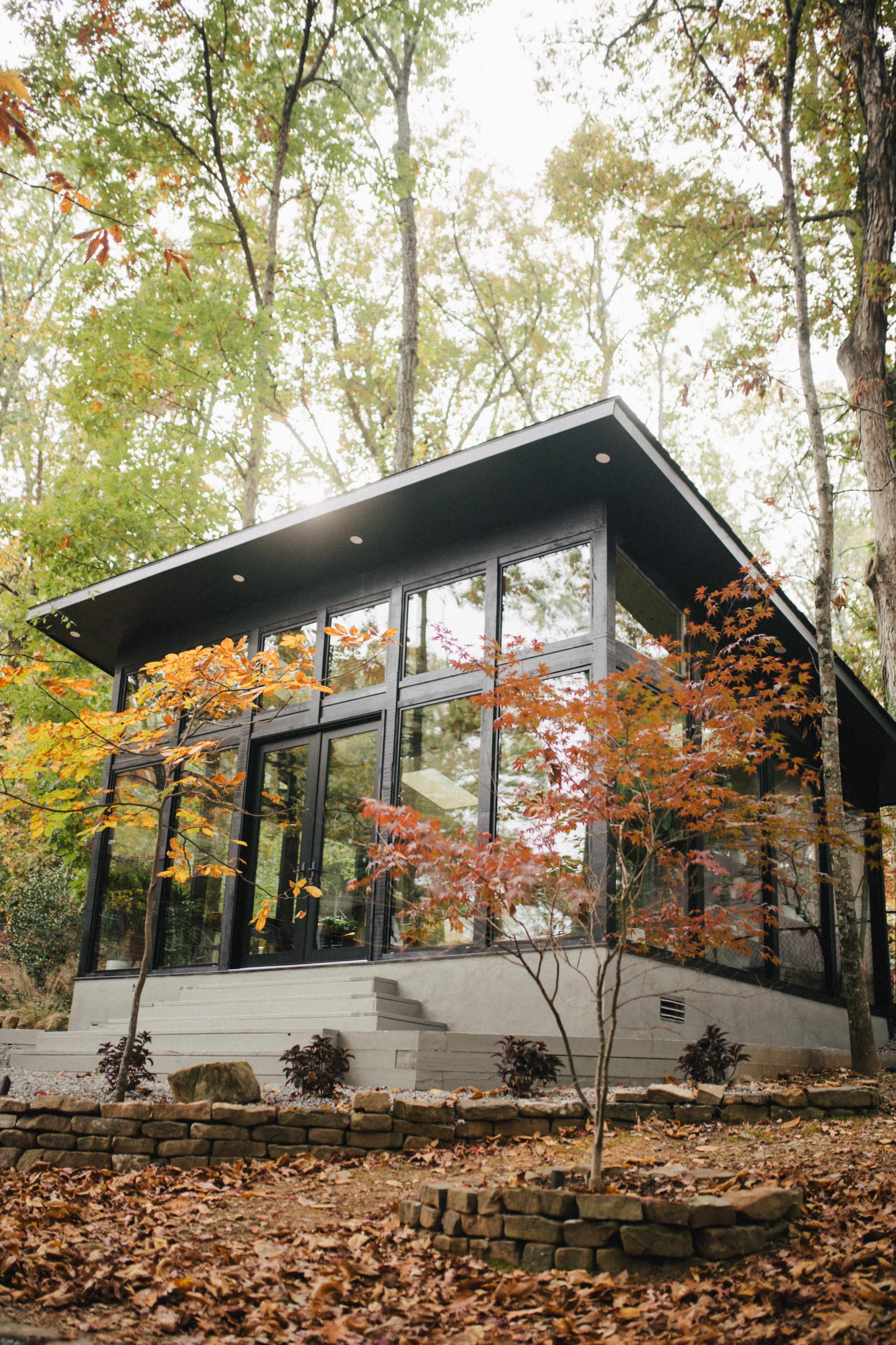 A modern, glass-fronted house is surrounded by trees with autumn foliage, set in a landscaped area with stone pathways.