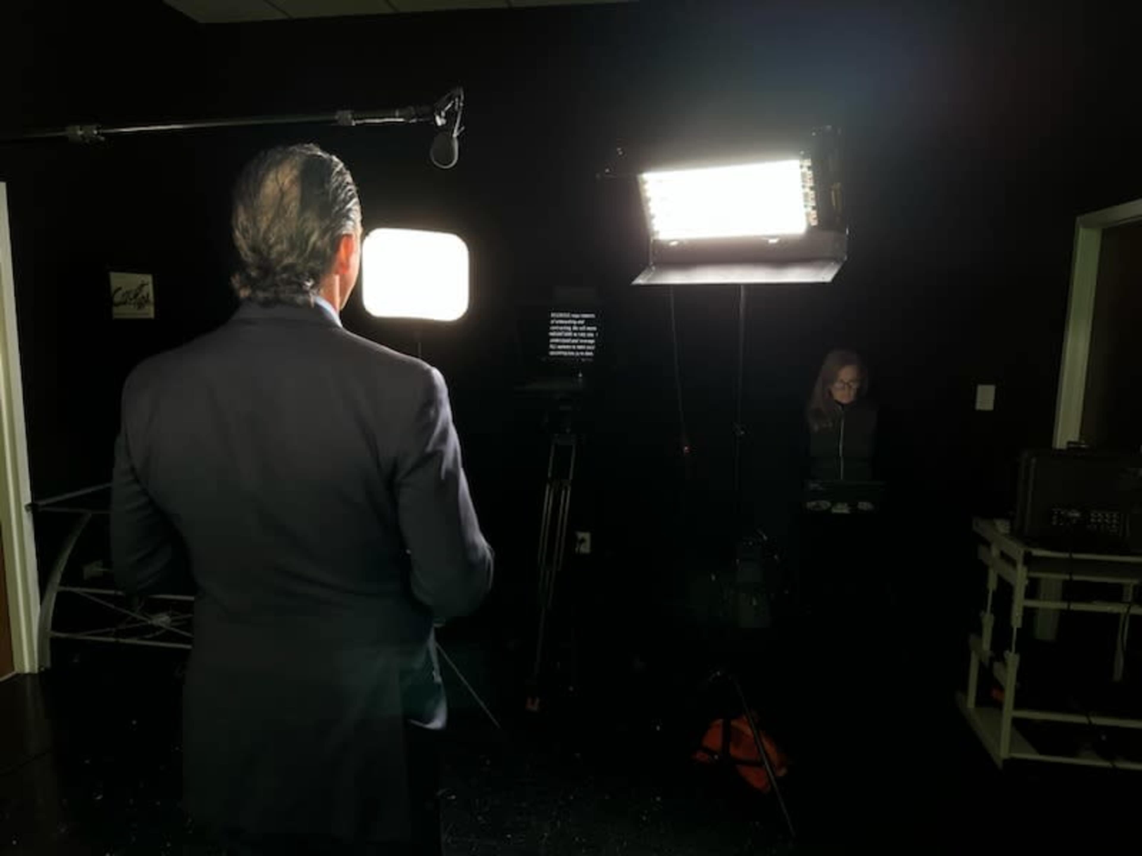 A man in a suit stands in front of bright studio lights while a woman is seated behind a control panel.
