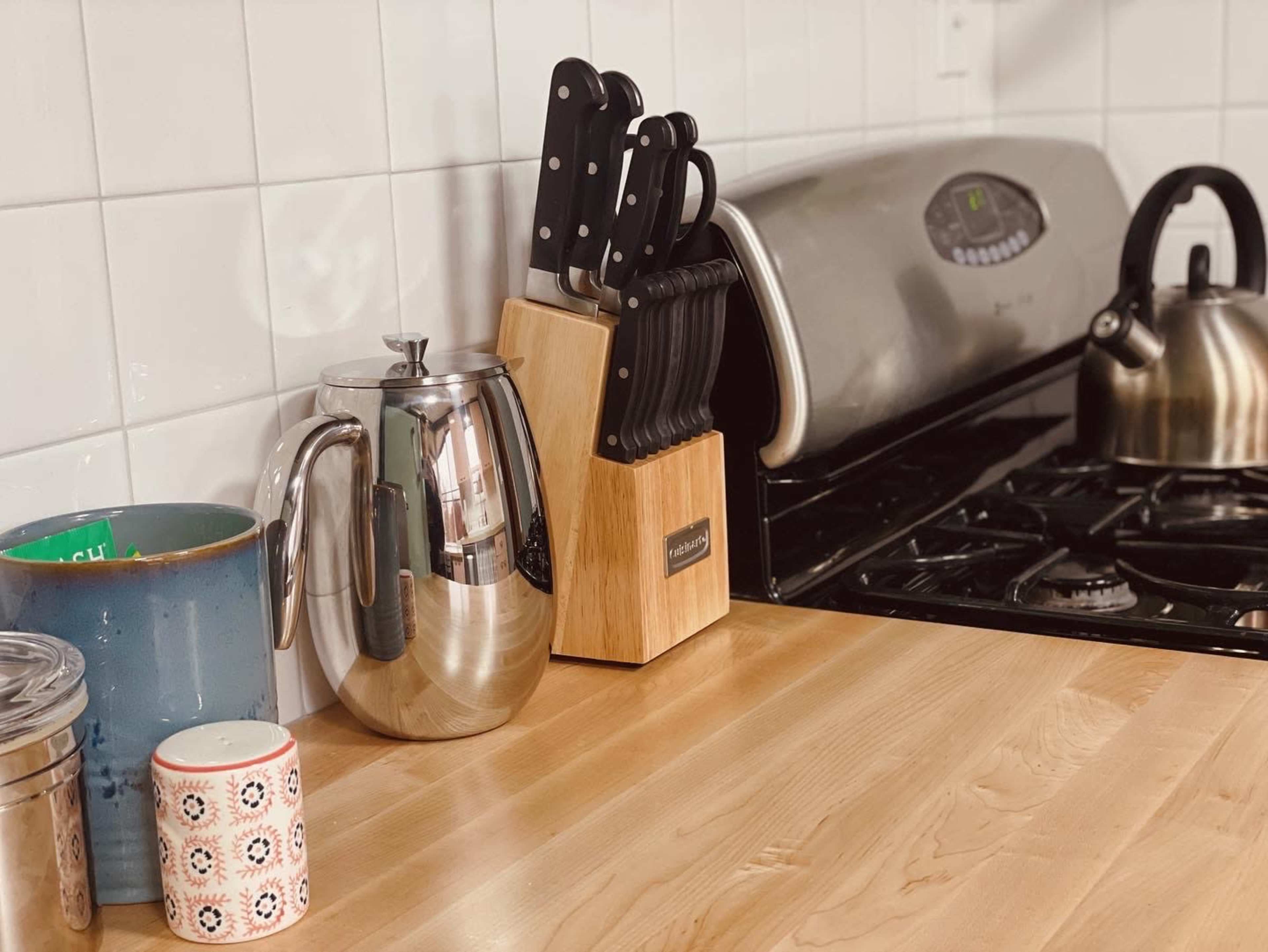 A stainless steel coffee pot, a wooden knife block with several knives, and a kettle are placed on a wooden kitchen countertop beside containers and a stovetop.