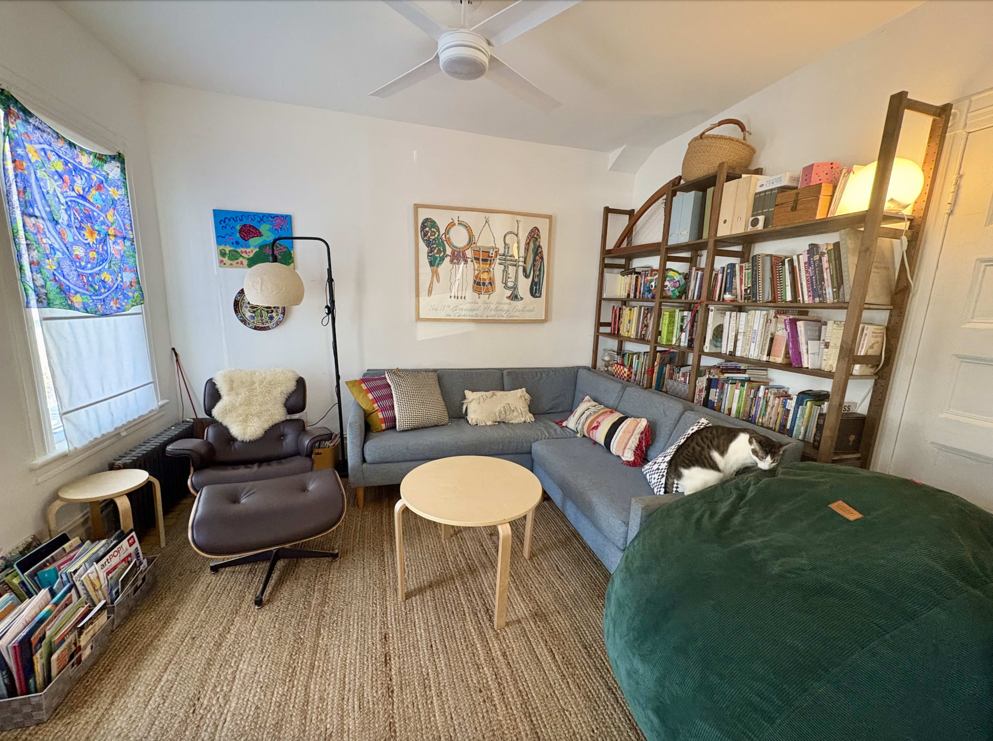 The image shows a cozy living room featuring a gray sectional sofa, a round coffee table, a large bean bag, a bookshelf filled with books, and decorative wall art.