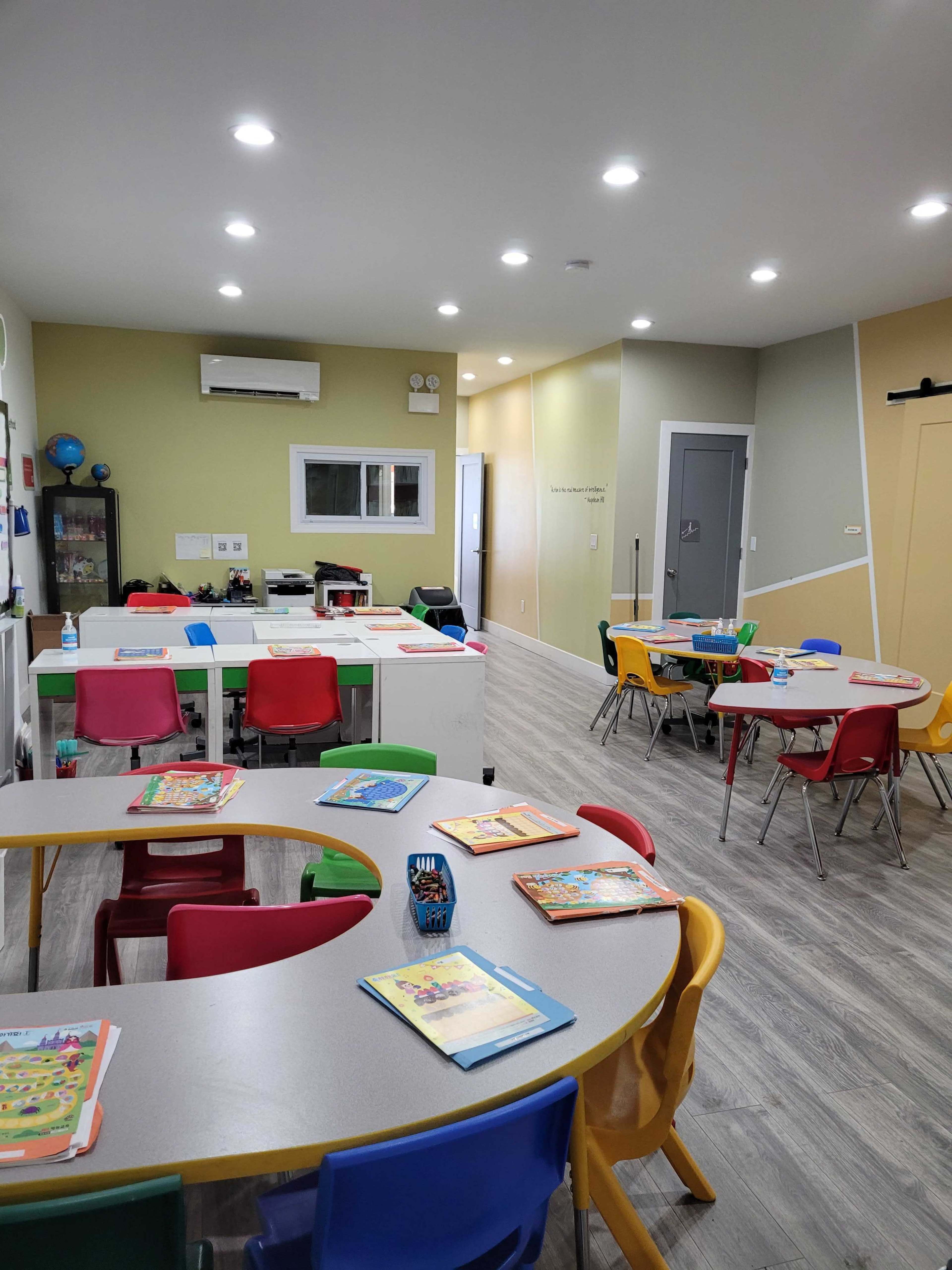 A brightly lit classroom features several tables with colorful chairs arranged, and books and materials laid out on the tables.