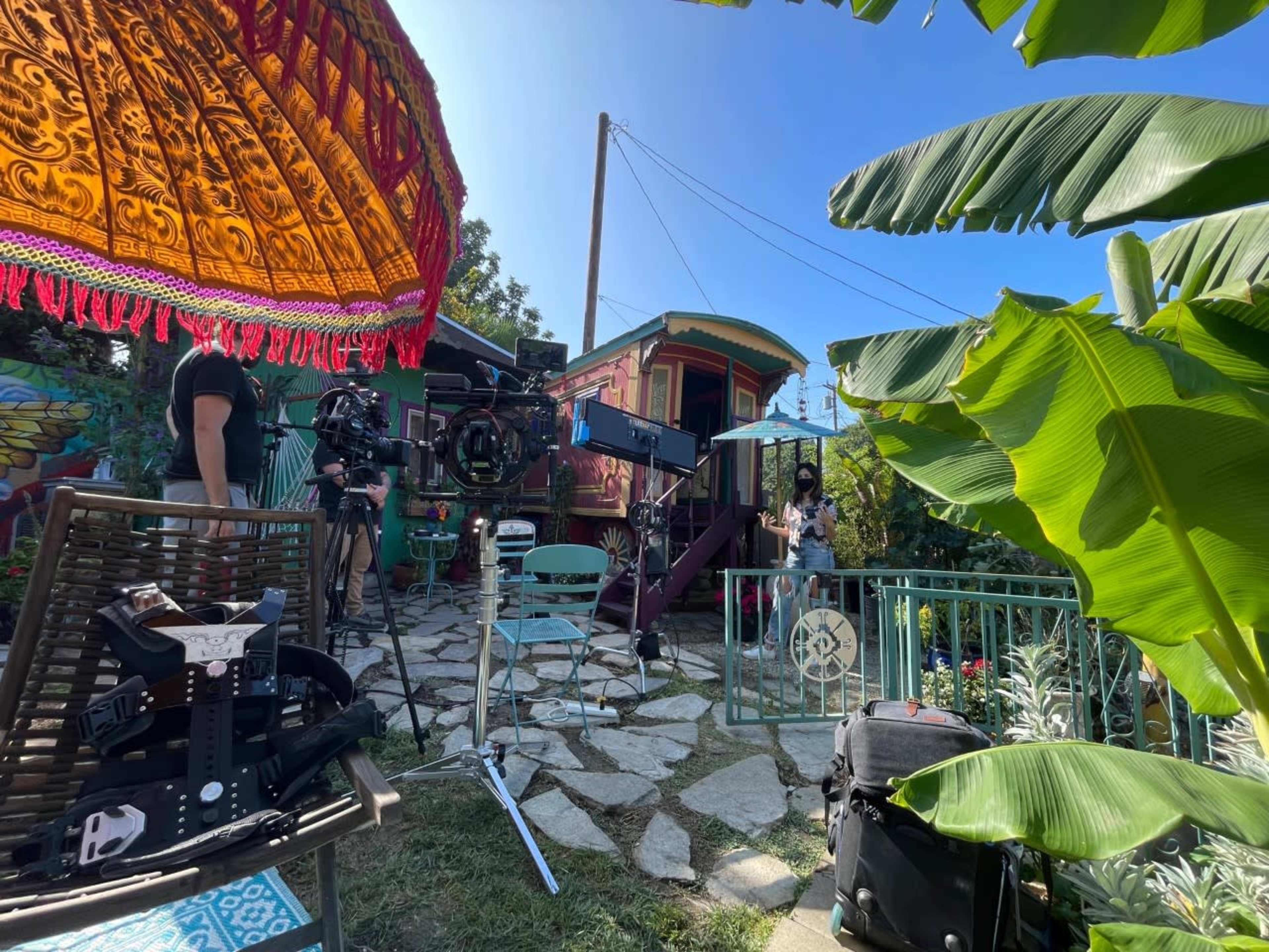 A film crew sets up equipment near a colorful trailer surrounded by lush greenery and vibrant decor.