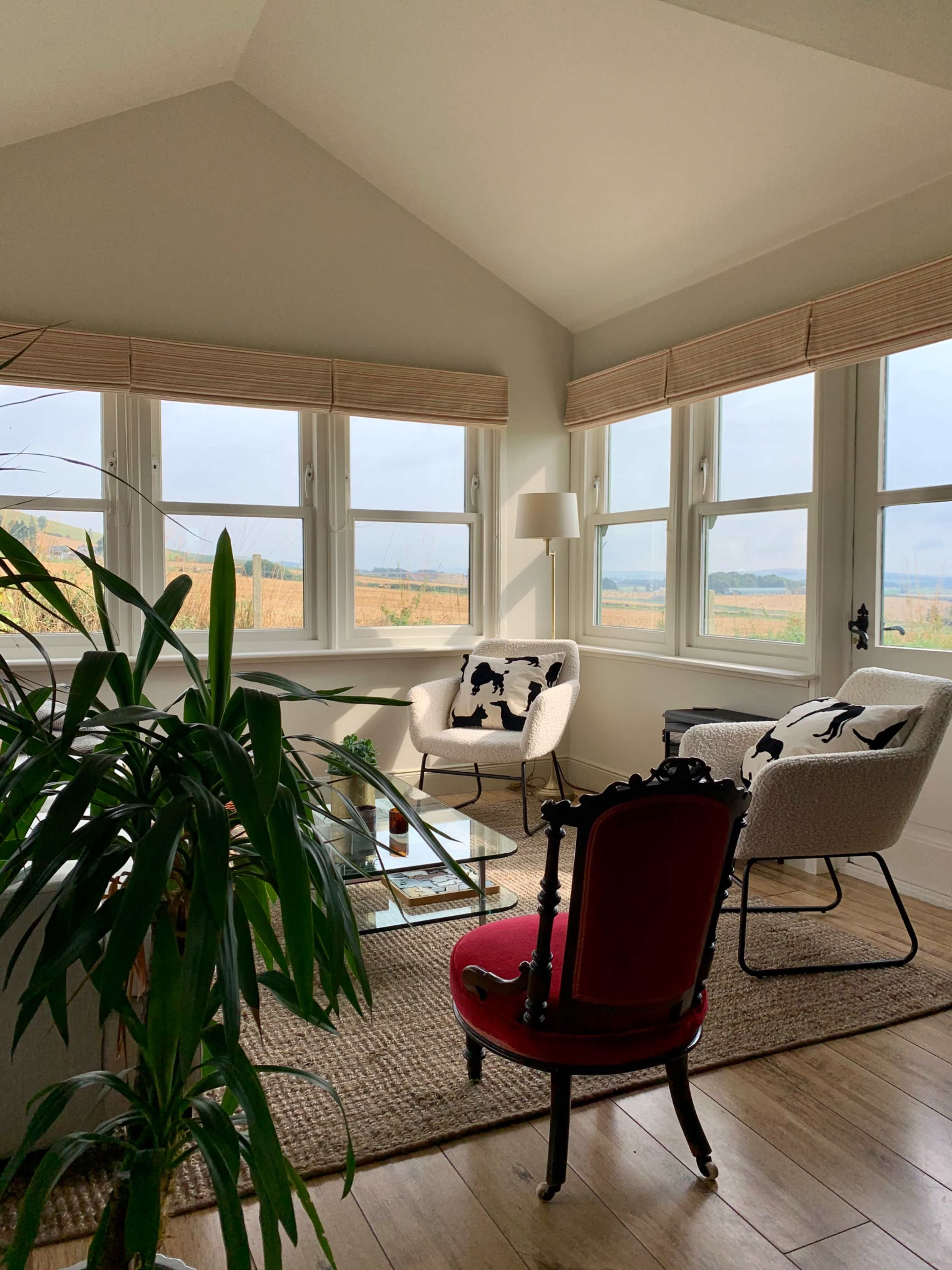 A bright sunroom features two white chairs, a glass coffee table, and large windows offering a view of a field.