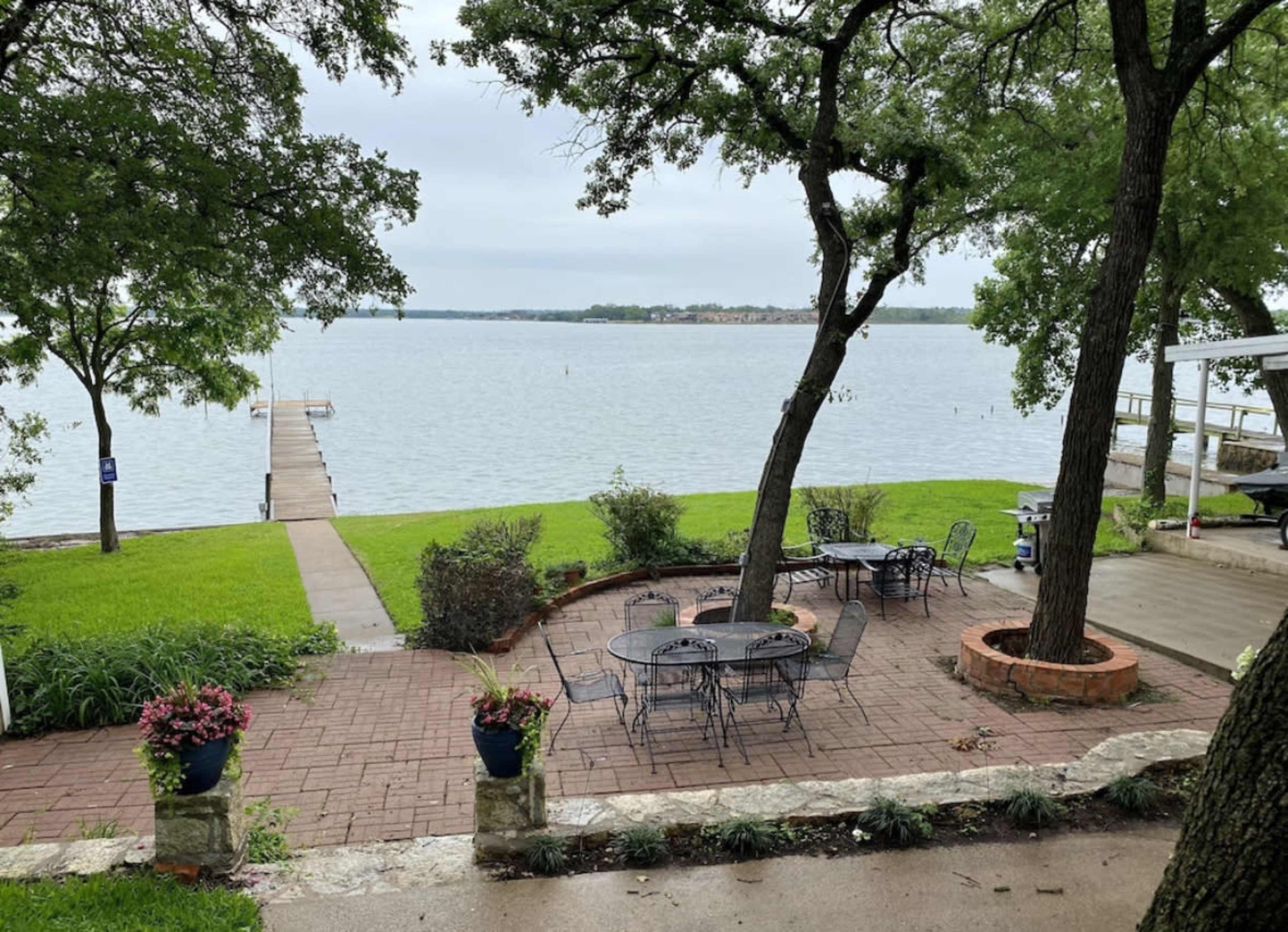 A patio with metal tables and chairs overlooks a calm lake, accompanied by a wooden dock extending into the water.