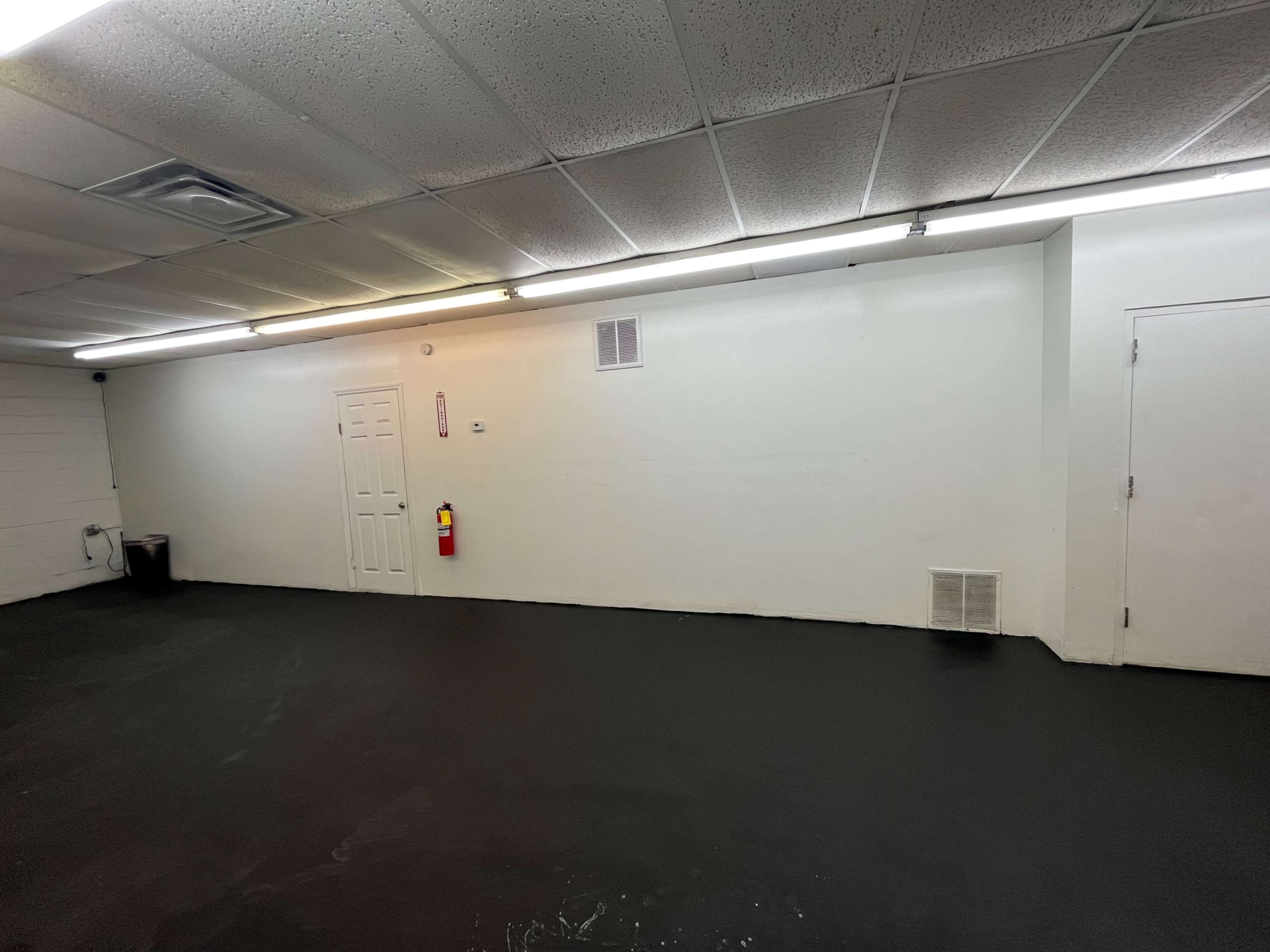 The image shows a large, empty room with a painted white wall, a closed door, and a fire extinguisher mounted on the wall.