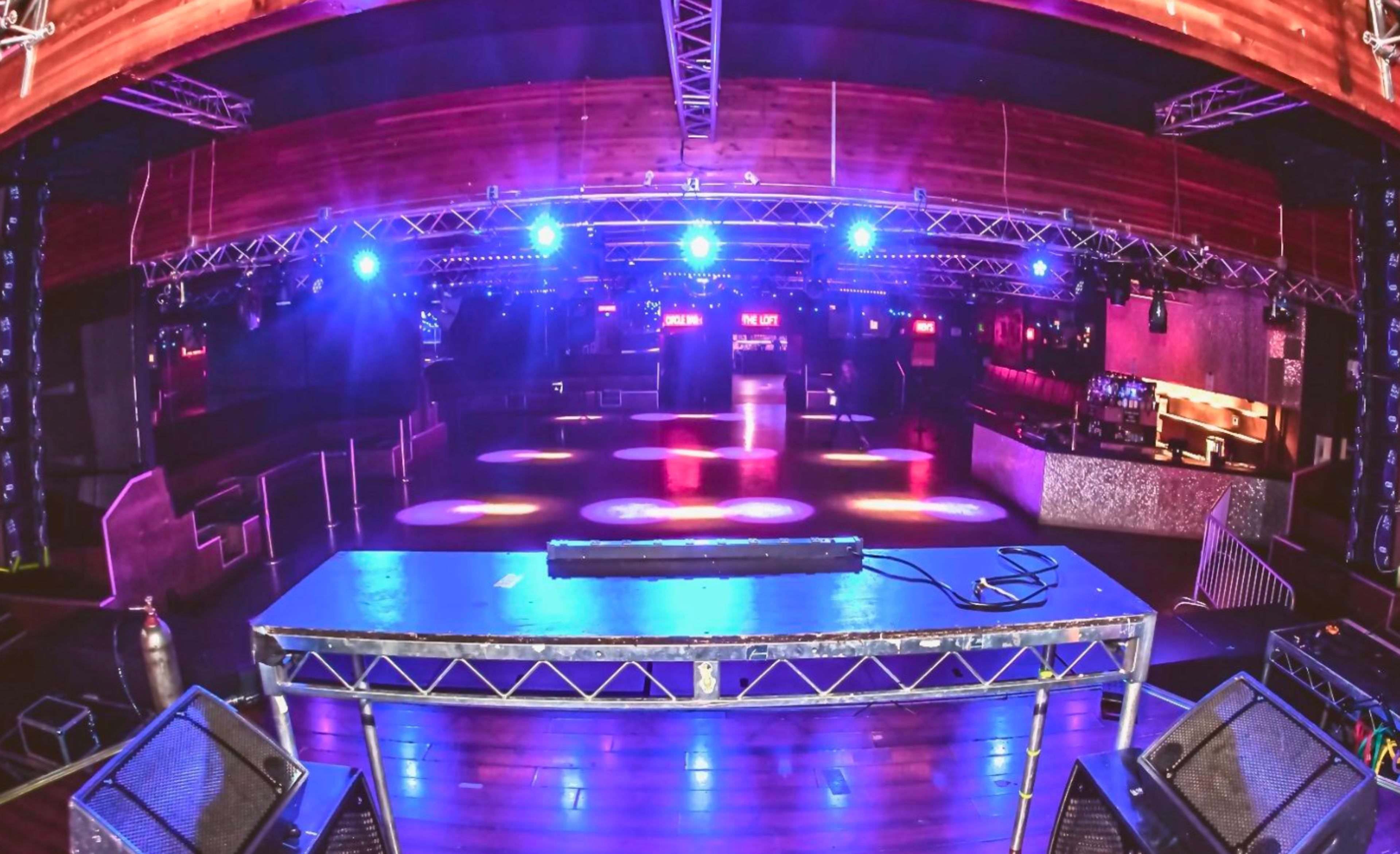 A spacious nightclub features a DJ booth in the foreground and vibrant colored lights illuminating the dance floor.