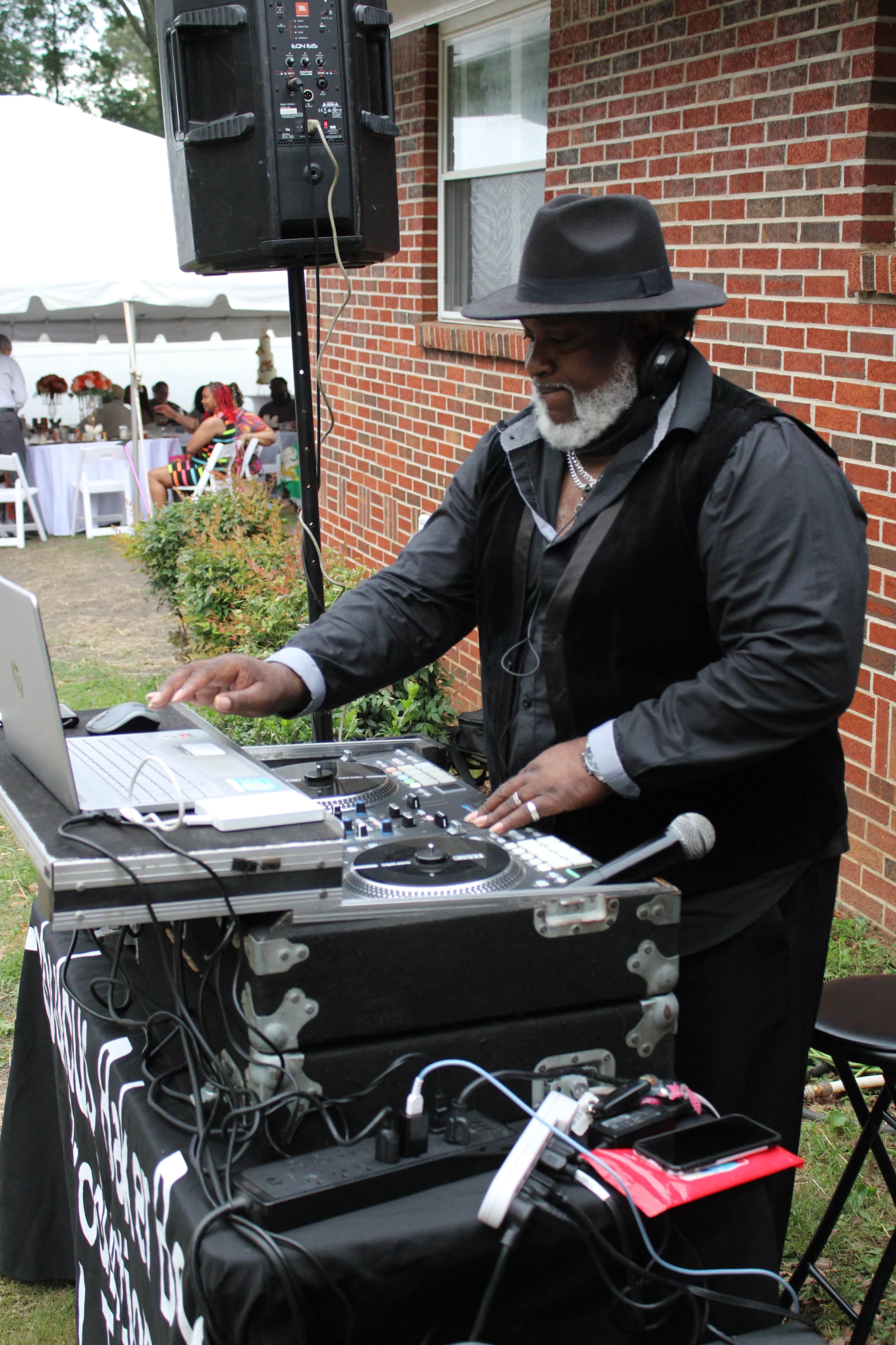 A DJ wearing a black vest and hat is mixing music on a laptop at an outdoor event with a tent in the background.