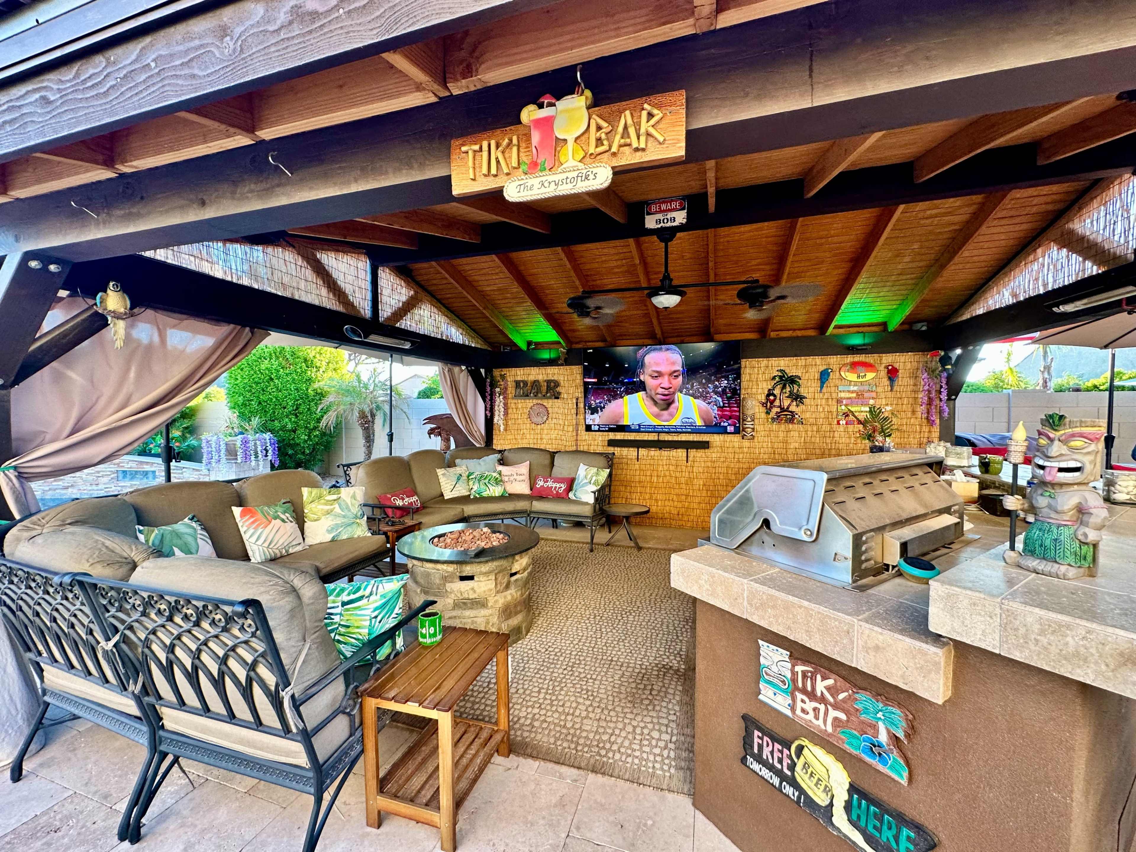 A tiki bar with a seating area, a television, and a grill is set up in a covered outdoor space.