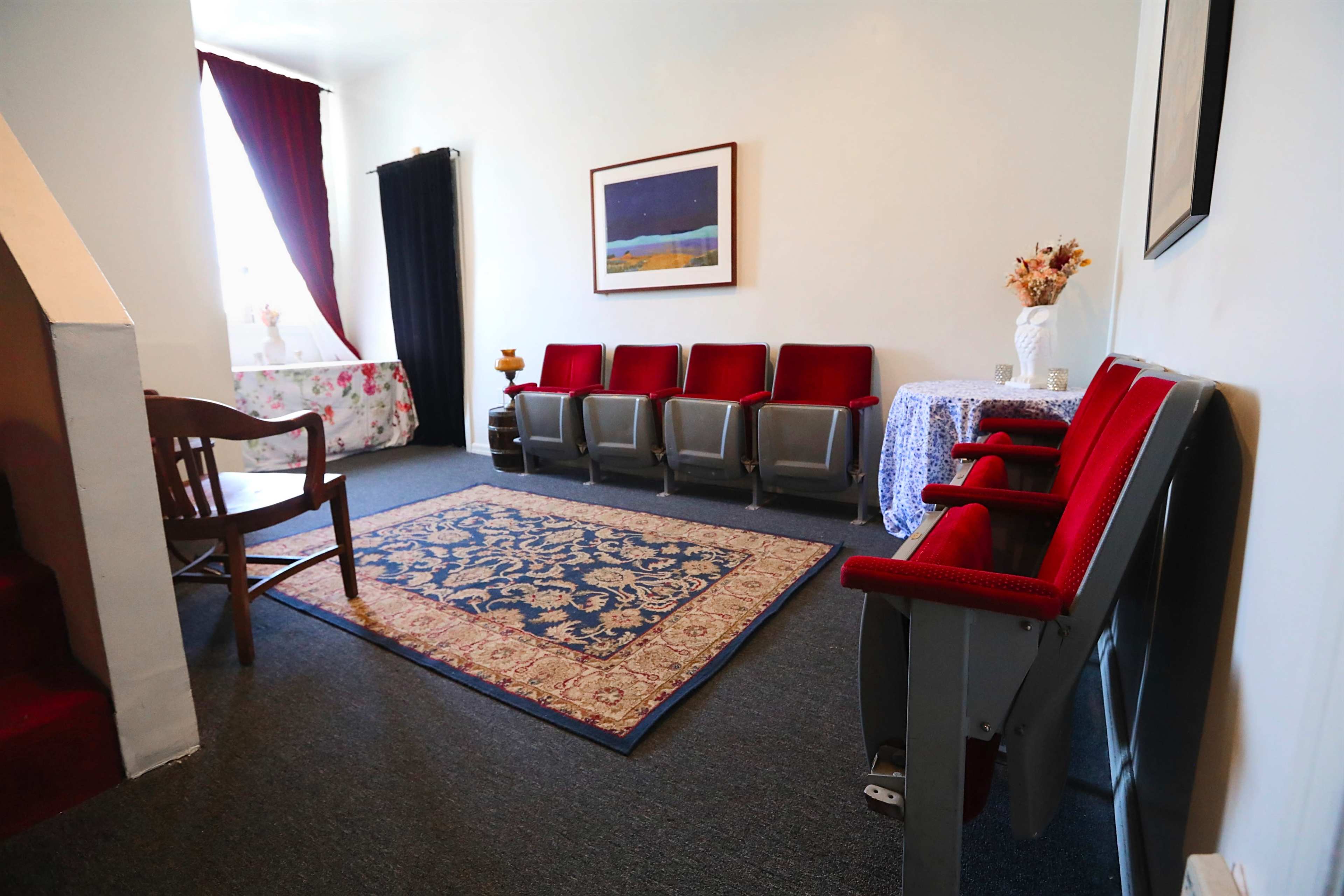 A small room with red chairs arranged in a row, a patterned area rug on the floor, and a table with a floral tablecloth.
