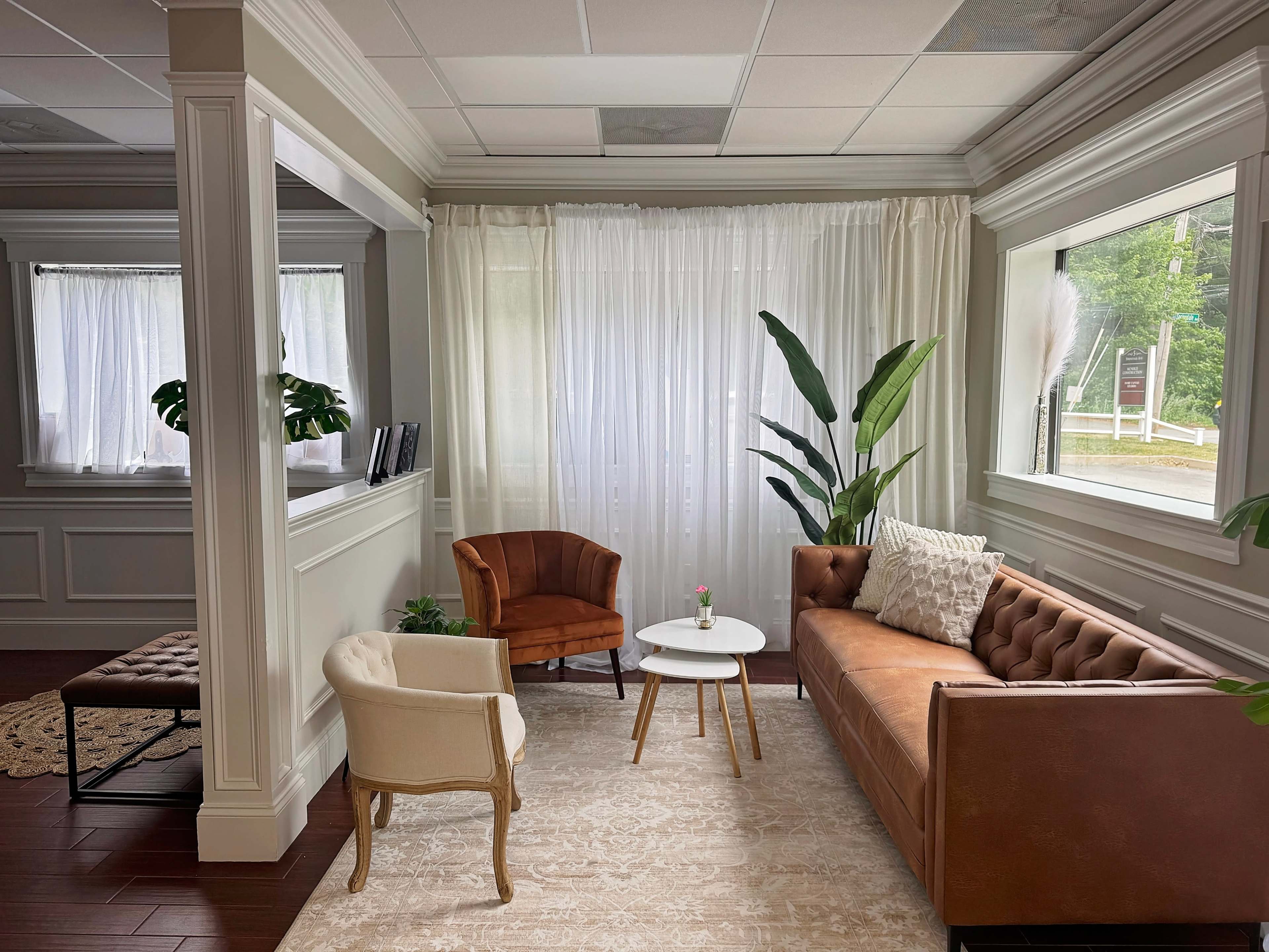 The image shows a well-lit waiting area featuring a brown leather sofa, a small round table, and two armchairs, with large windows draped in white curtains.
