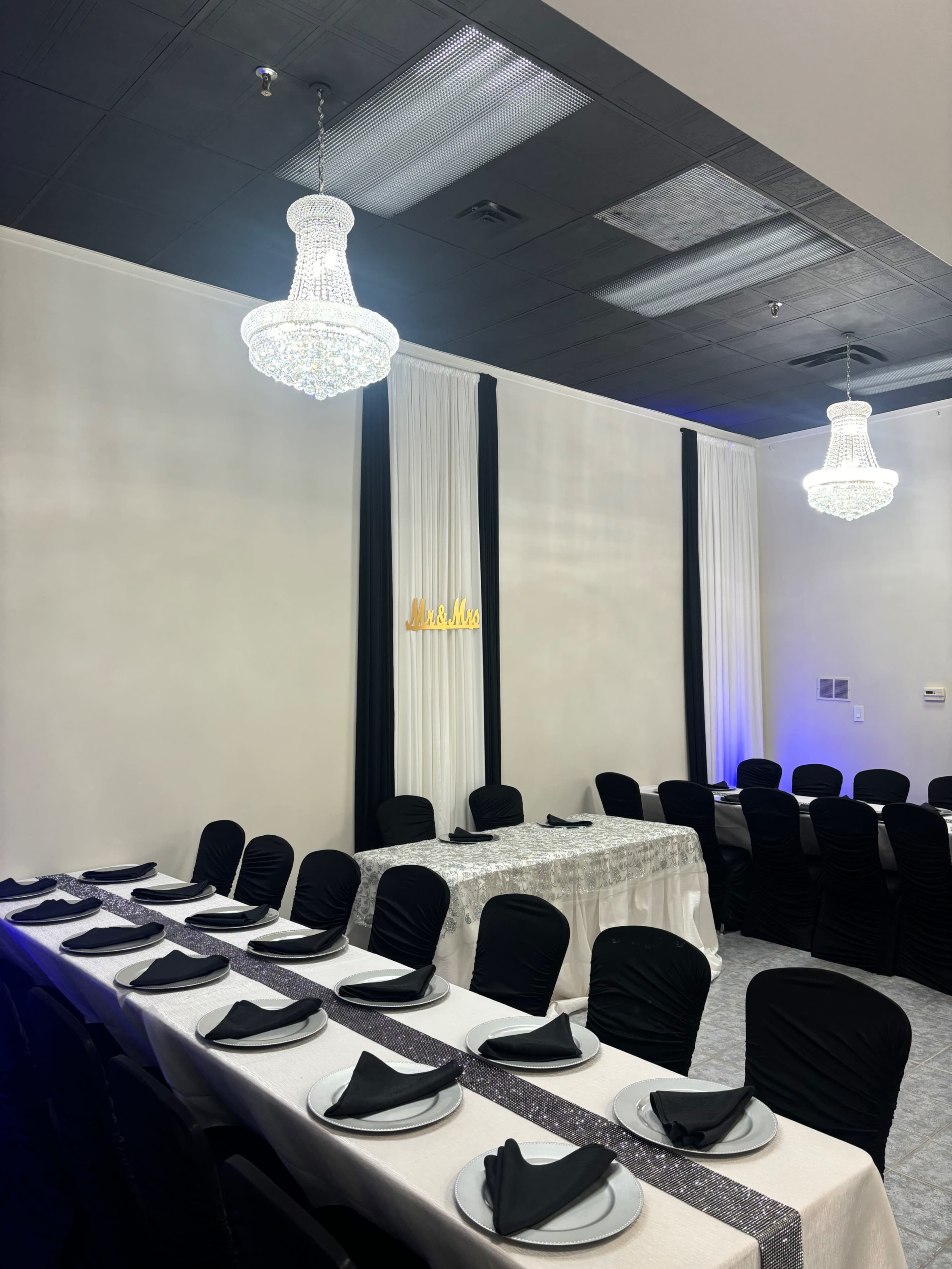 A dining area is set up with a long table adorned with black chairs, white tablecloths, and elegant chandeliers hanging from the ceiling.