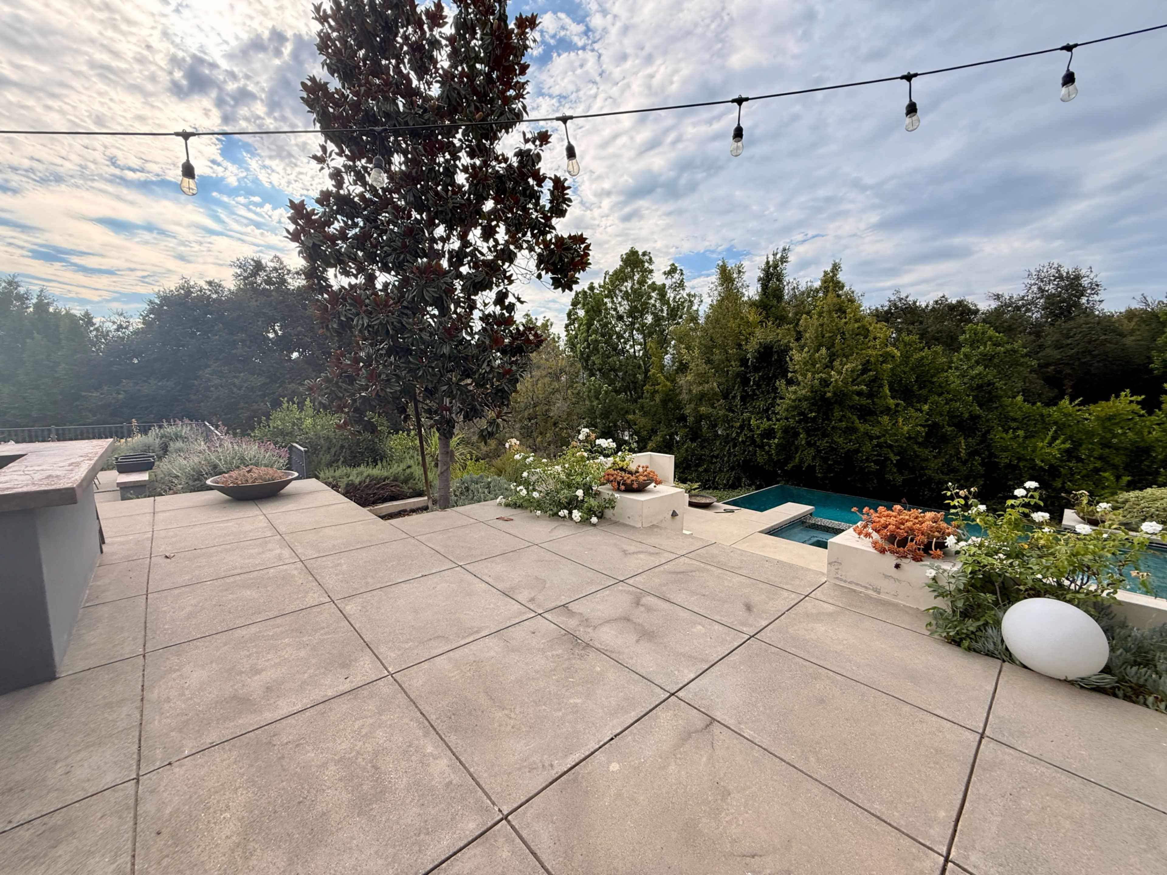 The image shows a patio area with potted plants, a swimming pool in the background, and a tree, under a cloudy sky with string lights overhead.