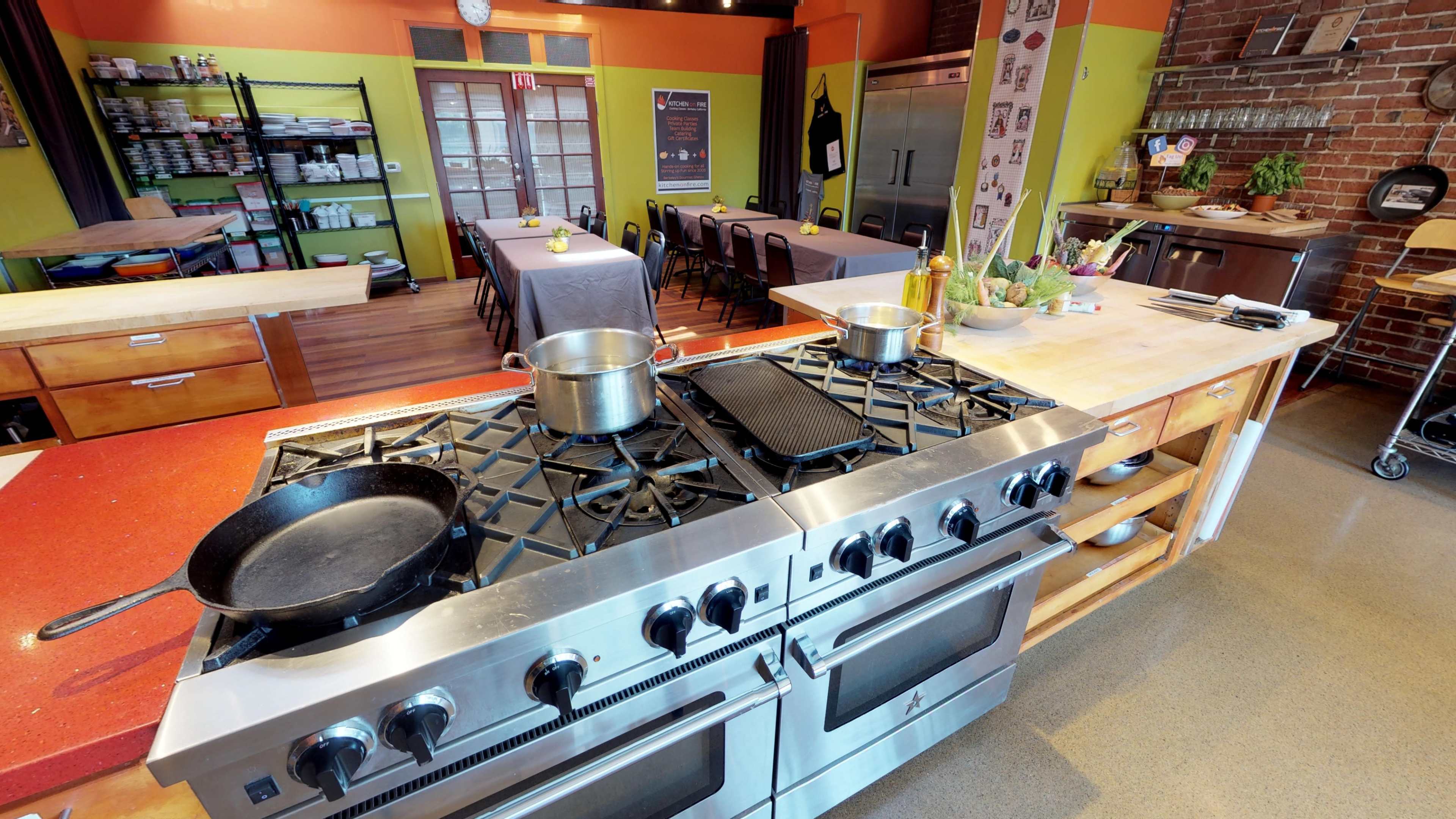 A commercial kitchen with a large stainless steel stove, pots on the burners, and a dining area featuring tables set for a meal.