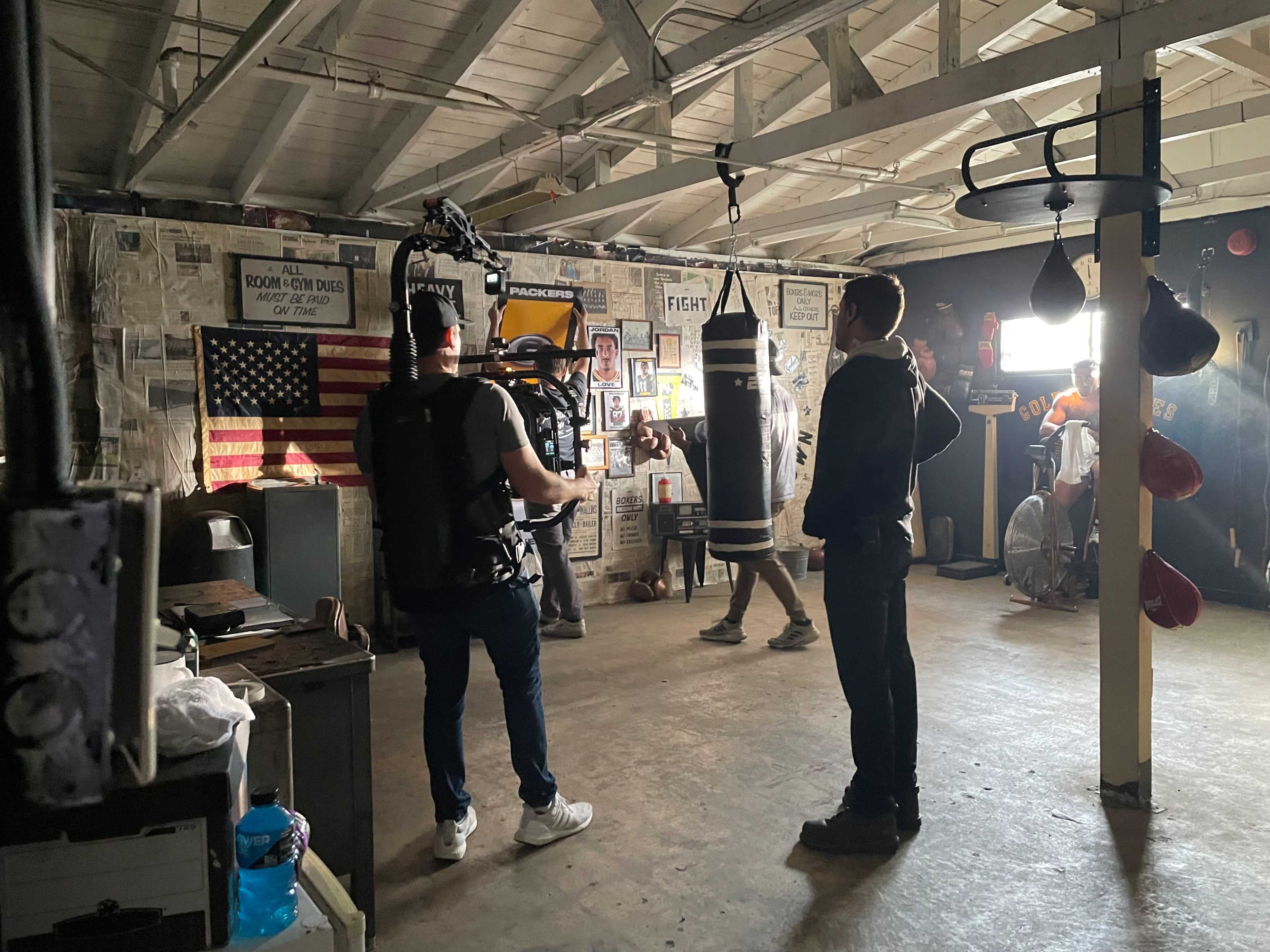 A film crew is shooting a scene in a boxing gym with a heavy bag and various boxing equipment visible on the walls.
