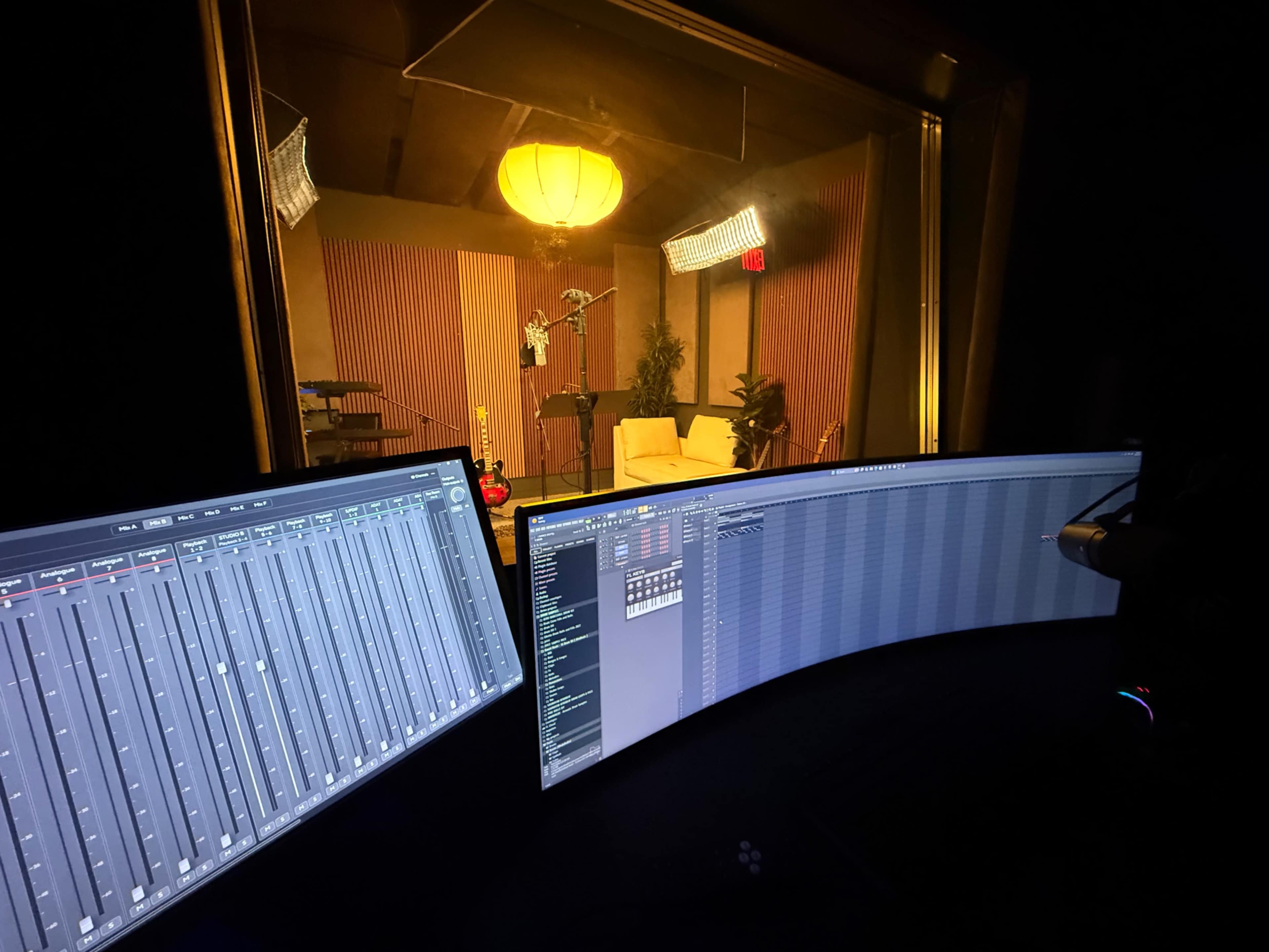 The image shows a recording studio view, featuring mixing consoles in the foreground and a well-lit sound booth in the background.