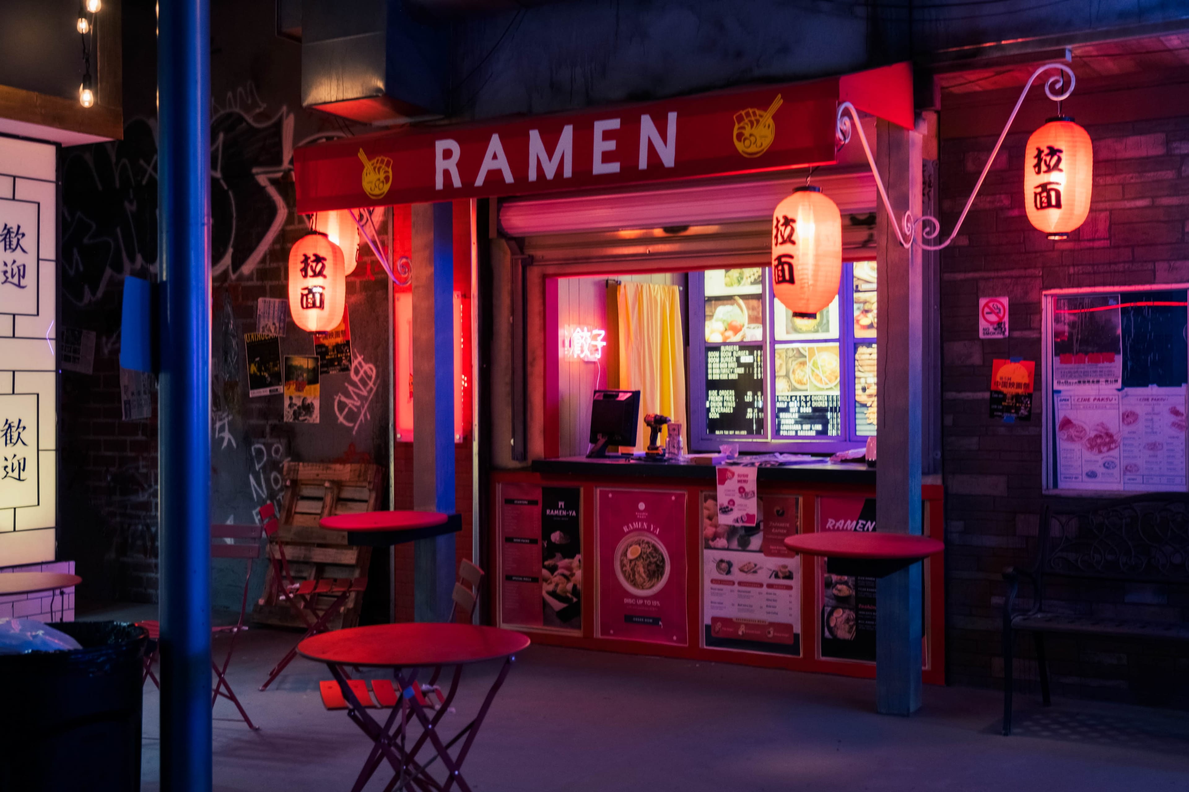 A ramen shop with red signage and lanterns is illuminated by colorful lights at night.